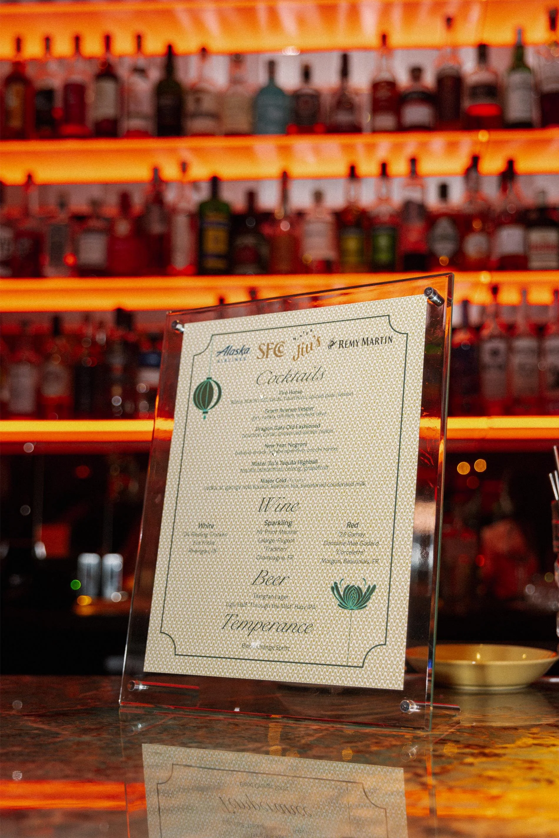A bar menu displayed on a stand with a neon-lit bar background featuring shelves of various liquor bottles.