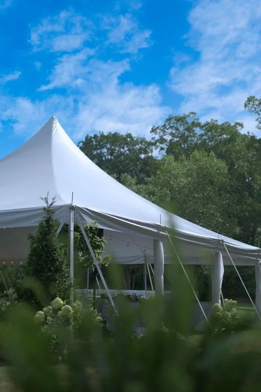 White event tent with pointed peak set up outdoors among trees and flowers under a partly cloudy blue sky.