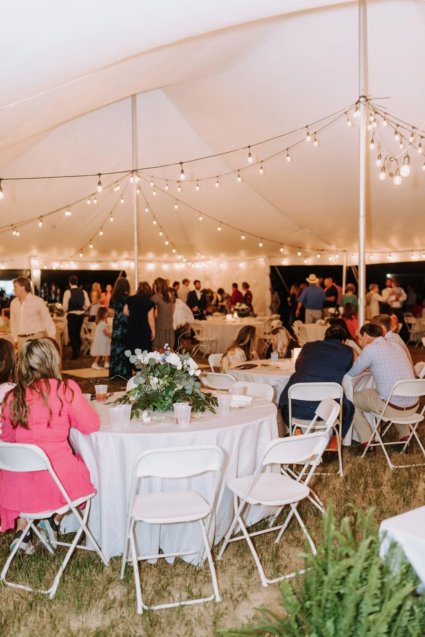 Guests seated at round tables with white tablecloths under a large white tent, decorated with string lights and floral centerpieces, at a nighttime event or celebration.