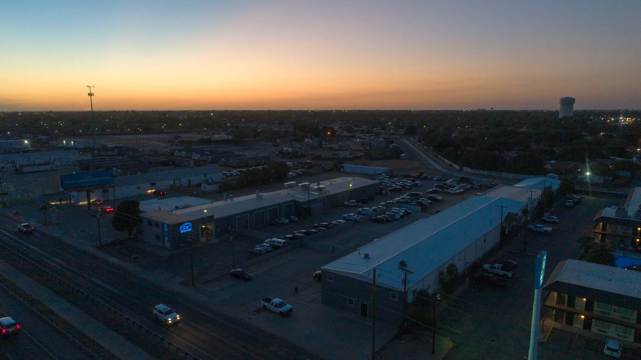 Nighttime aerial view of a commercial area with parking lots, buildings, and a street, under a sunset sky with some clouds on the horizon.