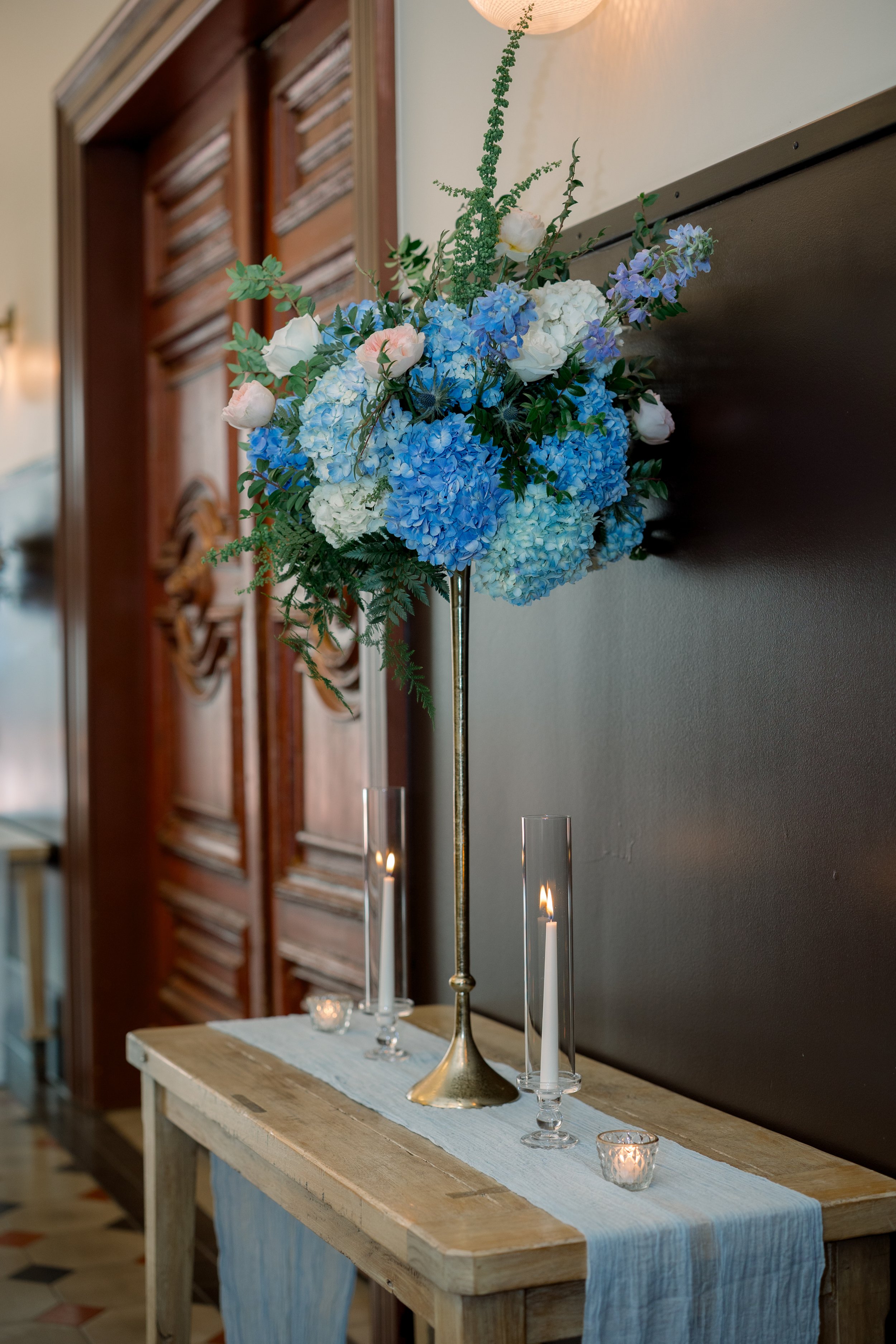 A tall, elegant flower arrangement with blue and white hydrangeas, white roses, and greenery on a wooden table with candles in glass holders, in an indoor setting with wooden wall decor.