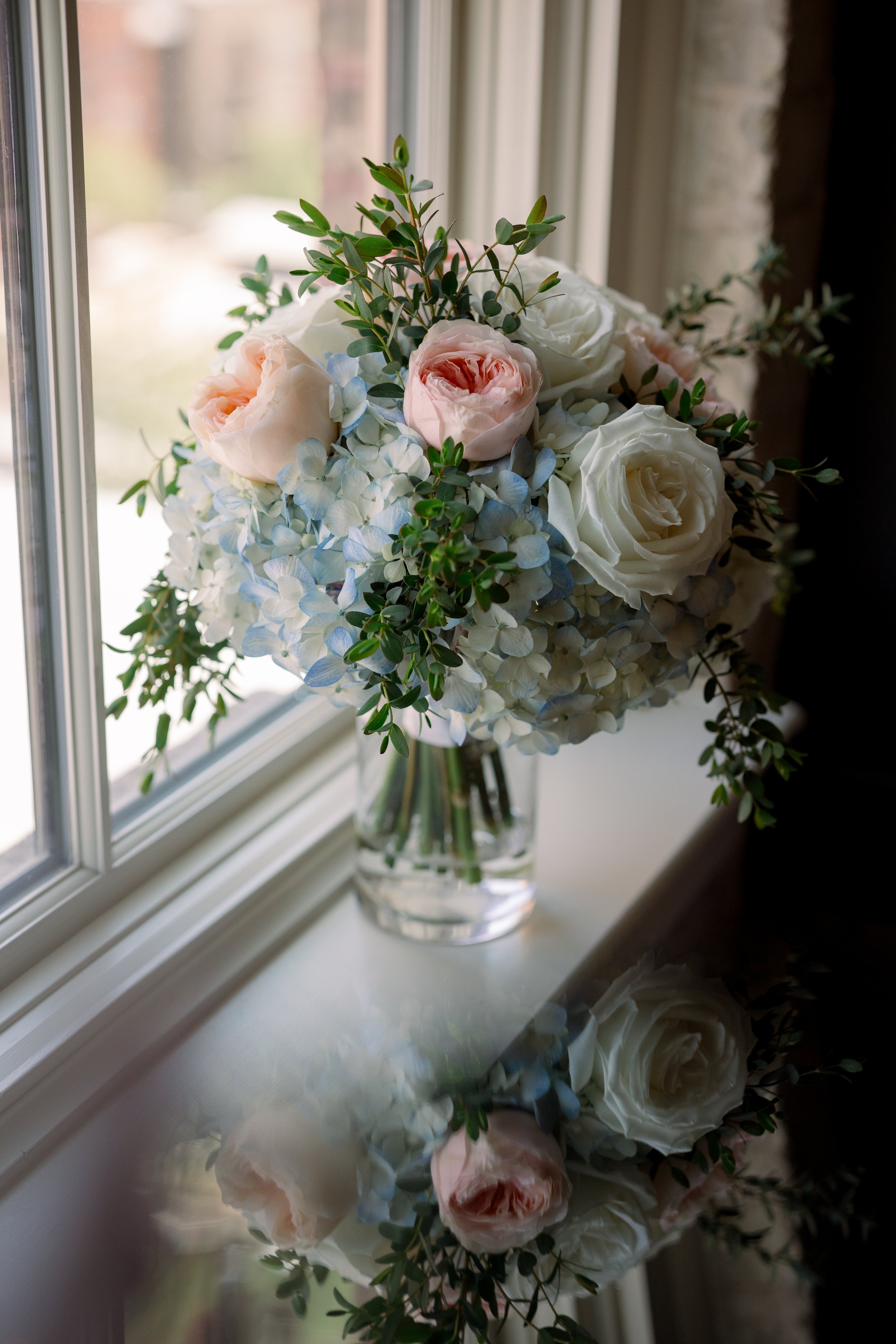 A bouquet of white and pale pink roses with blue hydrangeas and green foliage in a clear glass vase on a windowsill, with a reflection of the bouquet on the surface.