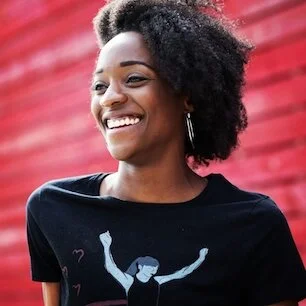 A smiling young woman with natural curly hair standing outdoors in front of a red wall.