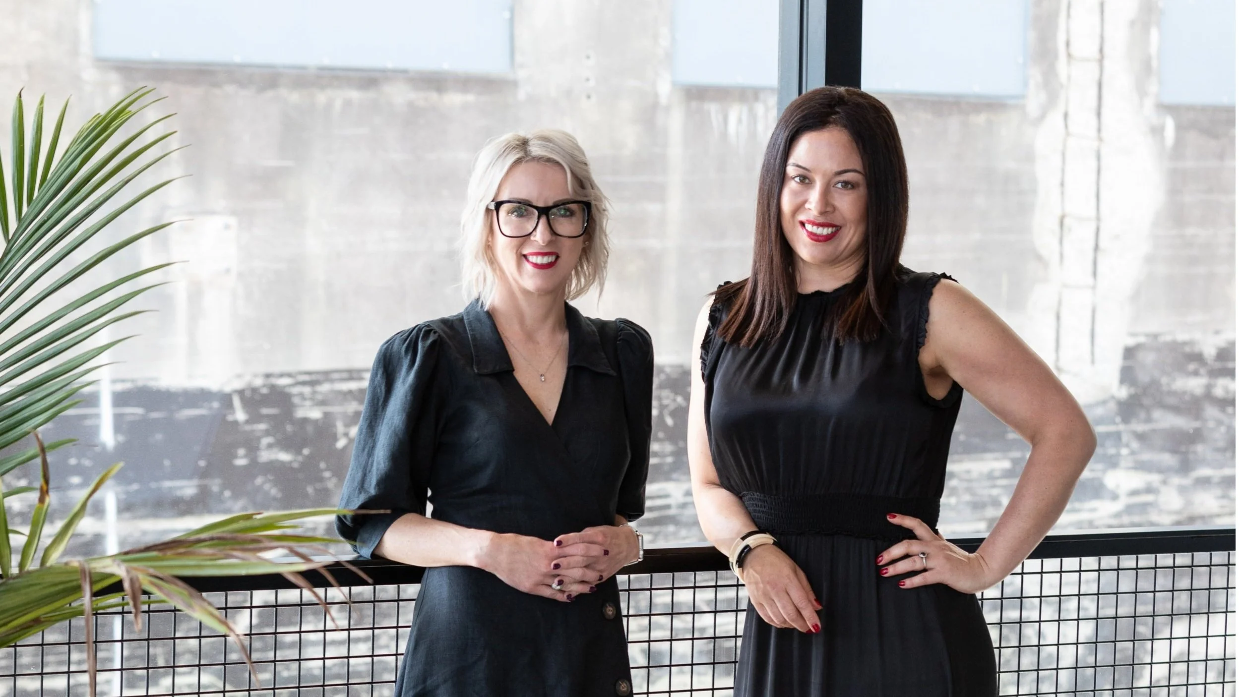 Two women standing indoors near a glass window with an industrial-style brick wall background. One woman has blonde hair, glasses, and is wearing a black jacket. The other woman has dark brown hair and is wearing a black sleeveless dress. They are smiling and looking at the camera.