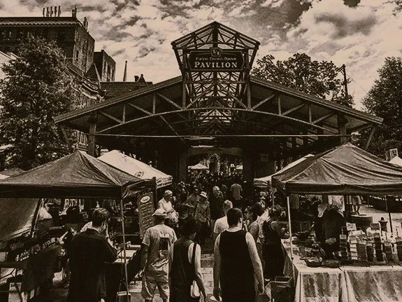 People walking through an outdoor market near Pavilion entrance with tents selling goods on either side, in front of a metal building structure with a sign reading 'Pavilion' at the top.