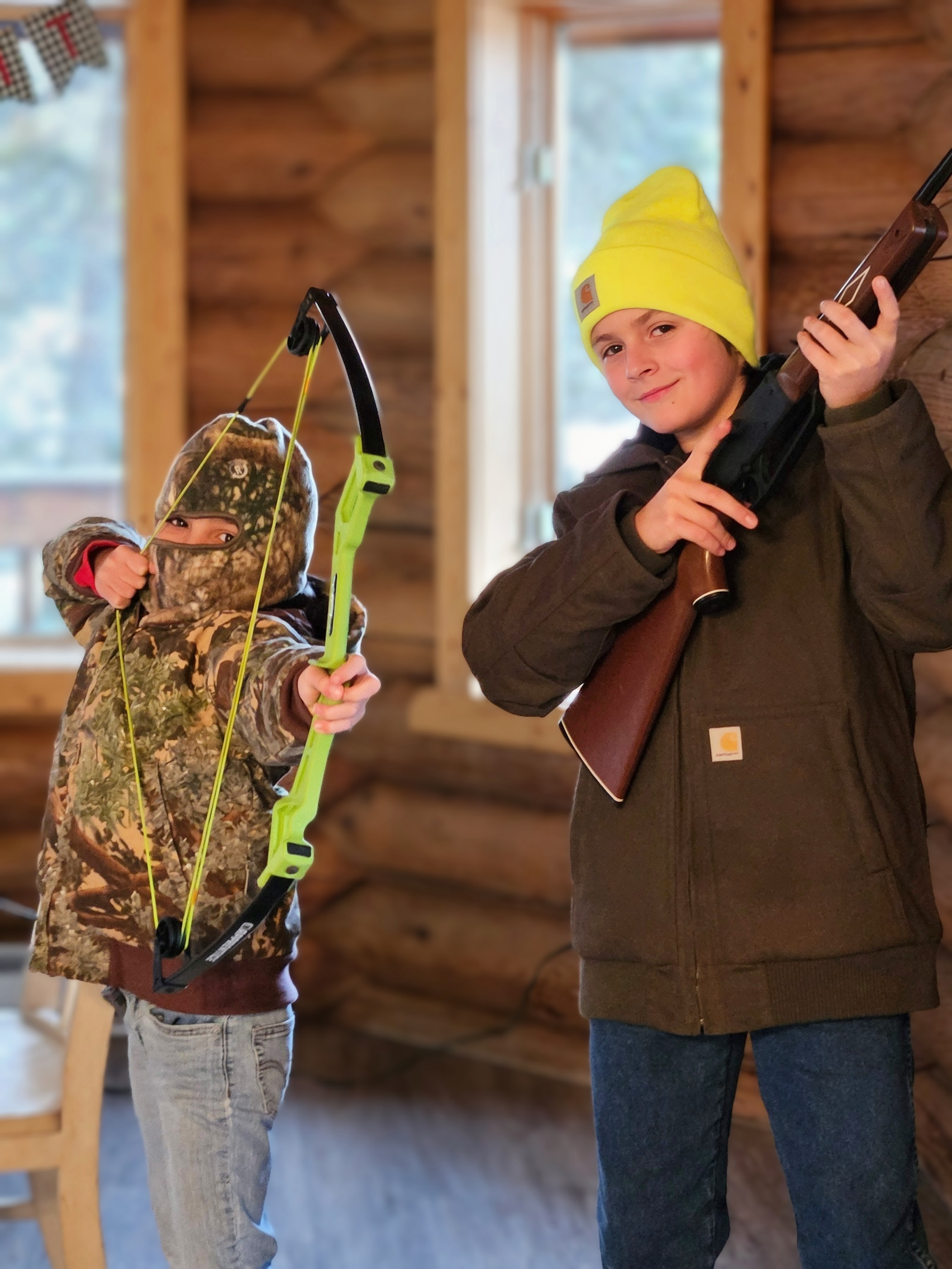 Two boys inside a log cabin, one holding a toy bow and arrow, the other holding a toy gun. The boy with the bow is dressed in camouflage and has a beanie, while the boy with the gun is wearing a bright yellow beanie and a brown jacket.