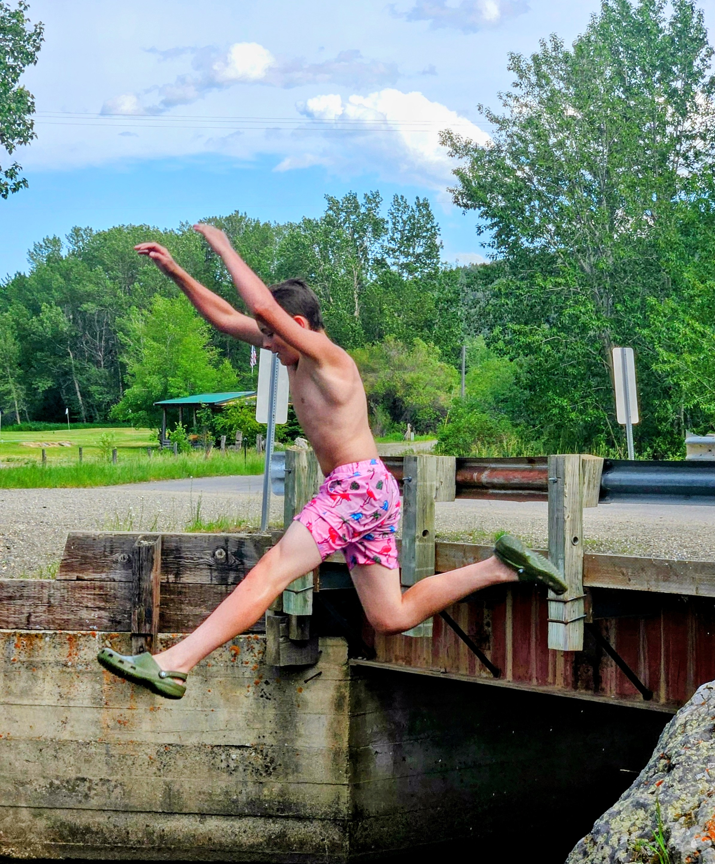 A young boy jumping into a body of water from a small wooden dock in a rural outdoor setting.