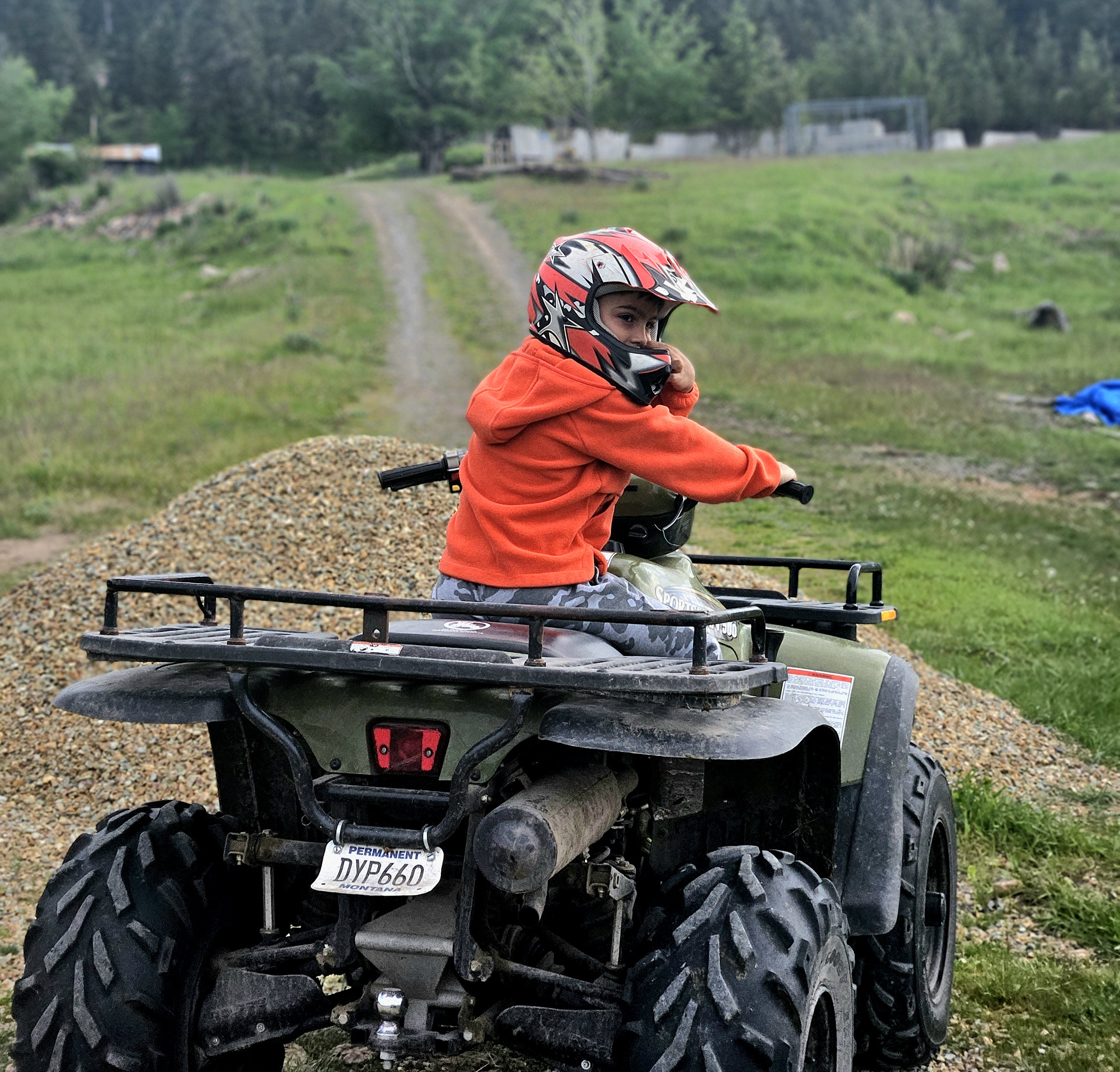 A young boy wearing a red helmet and orange hoodie sitting on a small green ATV in a grassy outdoor area with a dirt trail, trees, and some structures in the background.