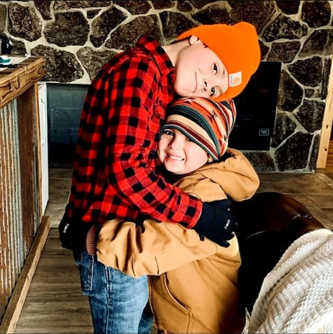Two young boys hugging indoors, dressed warmly with beanies and jackets, standing in front of a stone wall.
