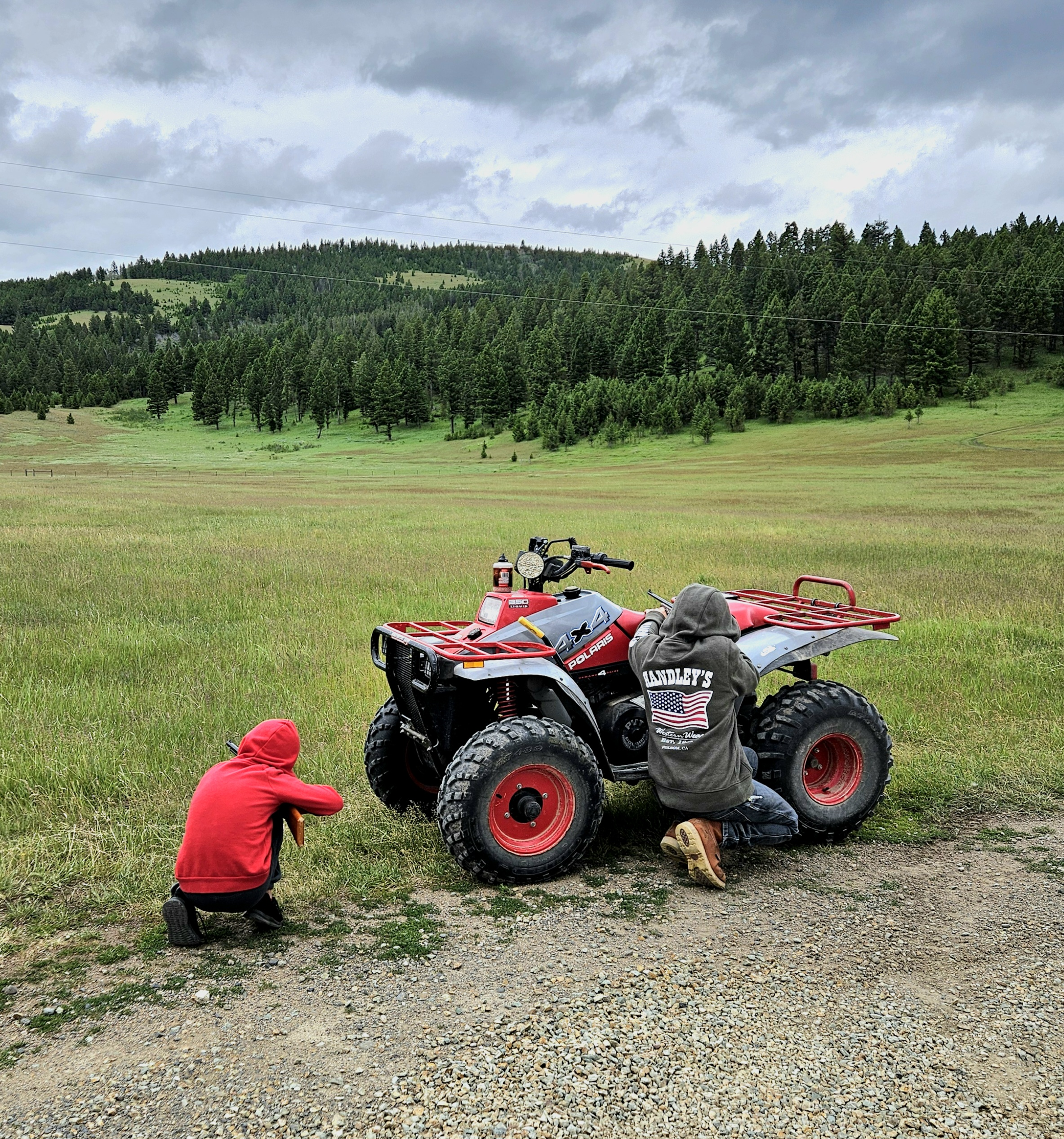 Two children in hoodies working on a red and black all-terrain vehicle (ATV) in a grassy field with forested hills and cloudy sky in the background.