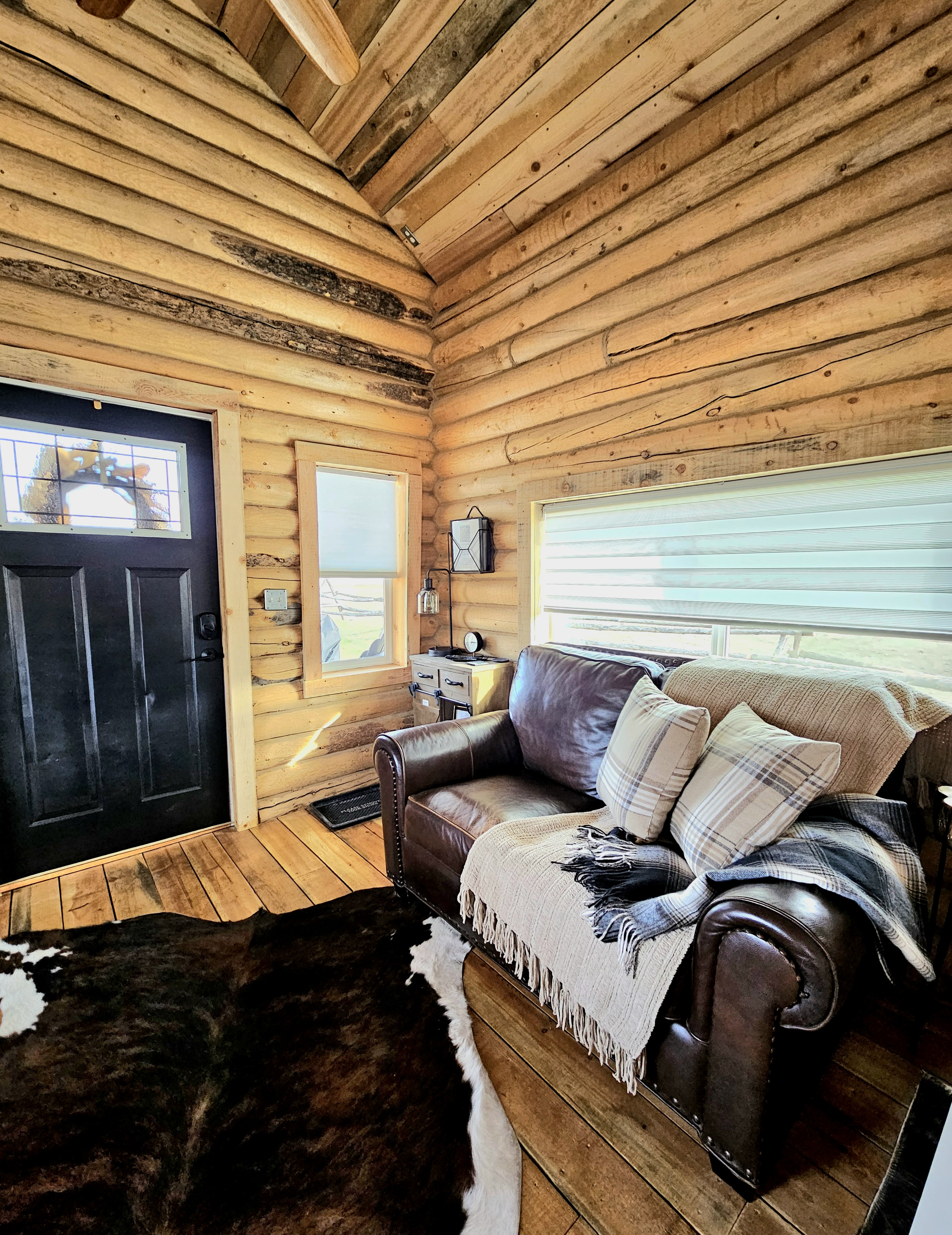 Interior of a rustic log cabin living room with wooden walls and floor, a black front door, a small window, a leather sofa with plaid pillows, a throw blanket, a cowhide rug, and a large window with blinds.