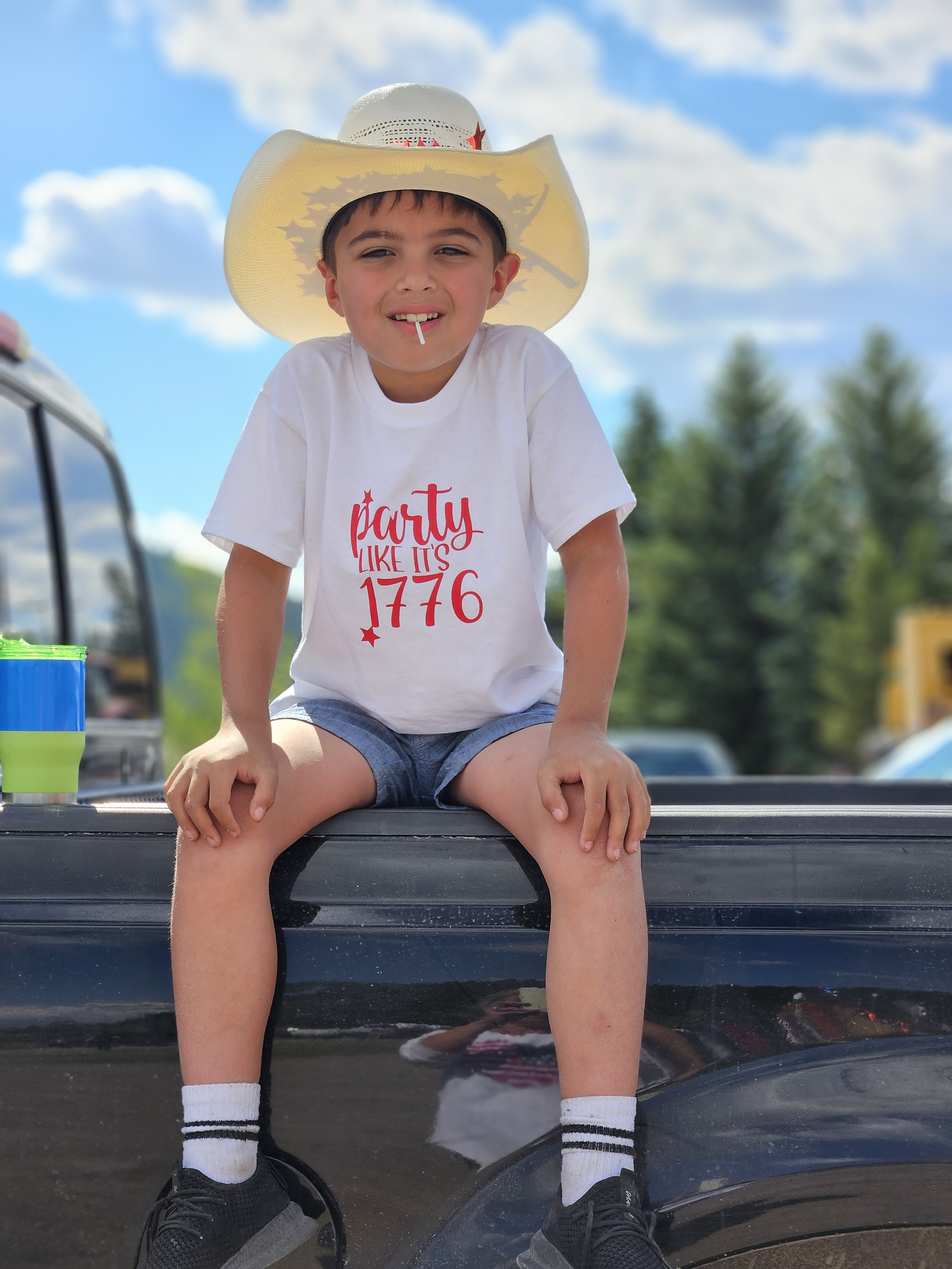 A young boy wearing a cowboy hat, white t-shirt with red text, shorts, and sneakers, sitting on the back of a truck during daytime with a blue sky and trees in the background.