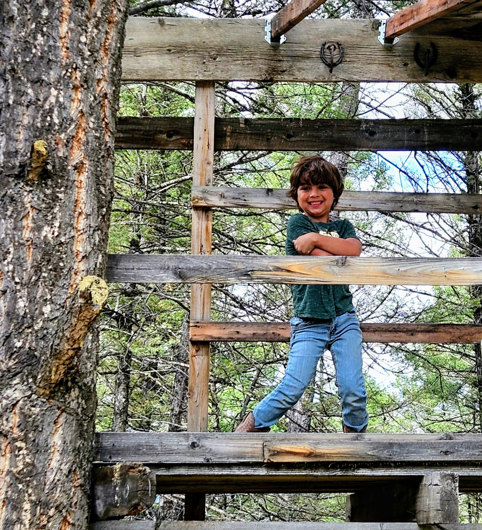 Young boy standing on a wooden treehouse platform, smiling with arms crossed, surrounded by trees.