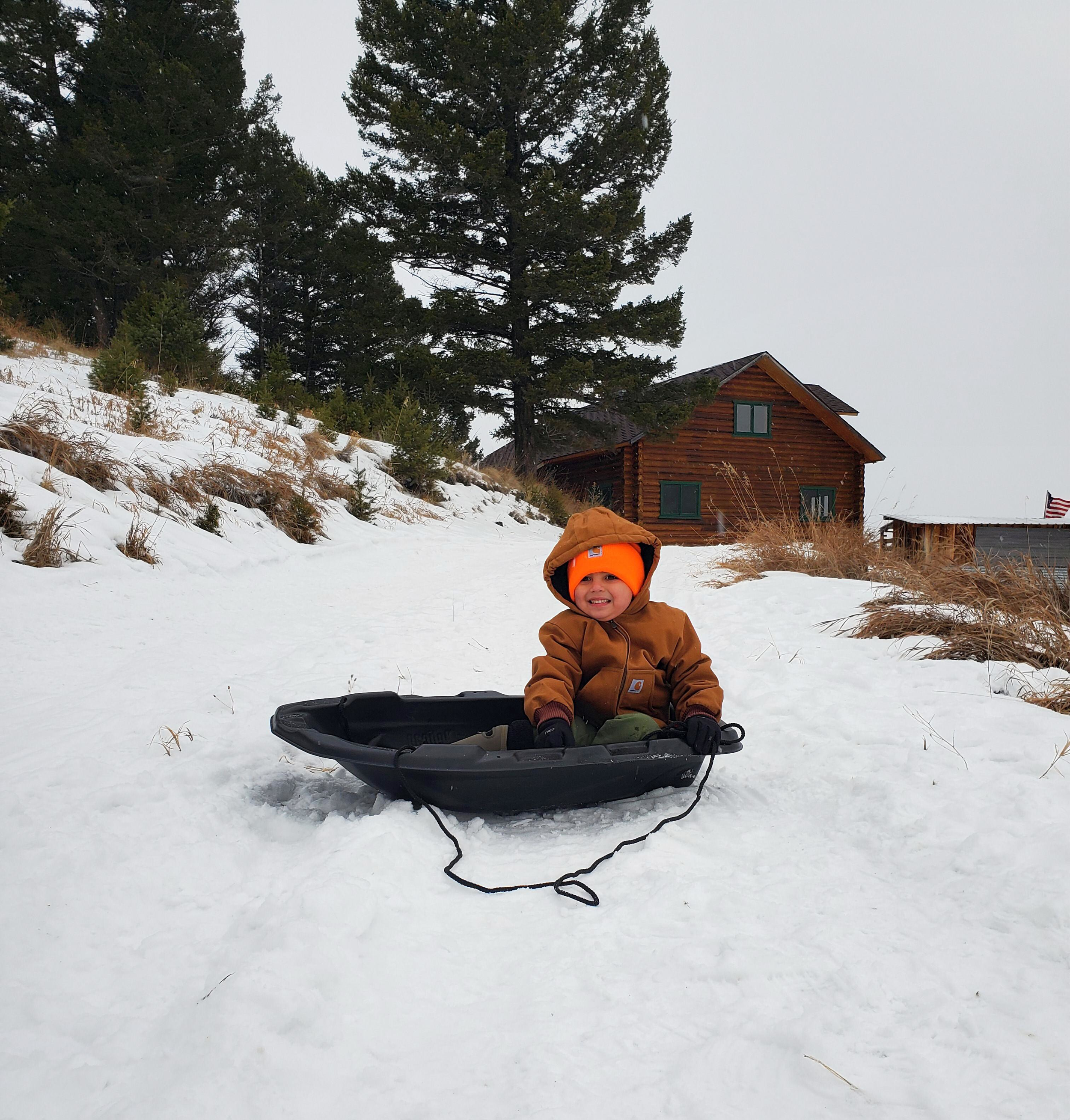 A young child bundled in a brown jacket and orange hat sitting in a black snow sled on a snow-covered path, with a log cabin and trees in the background.