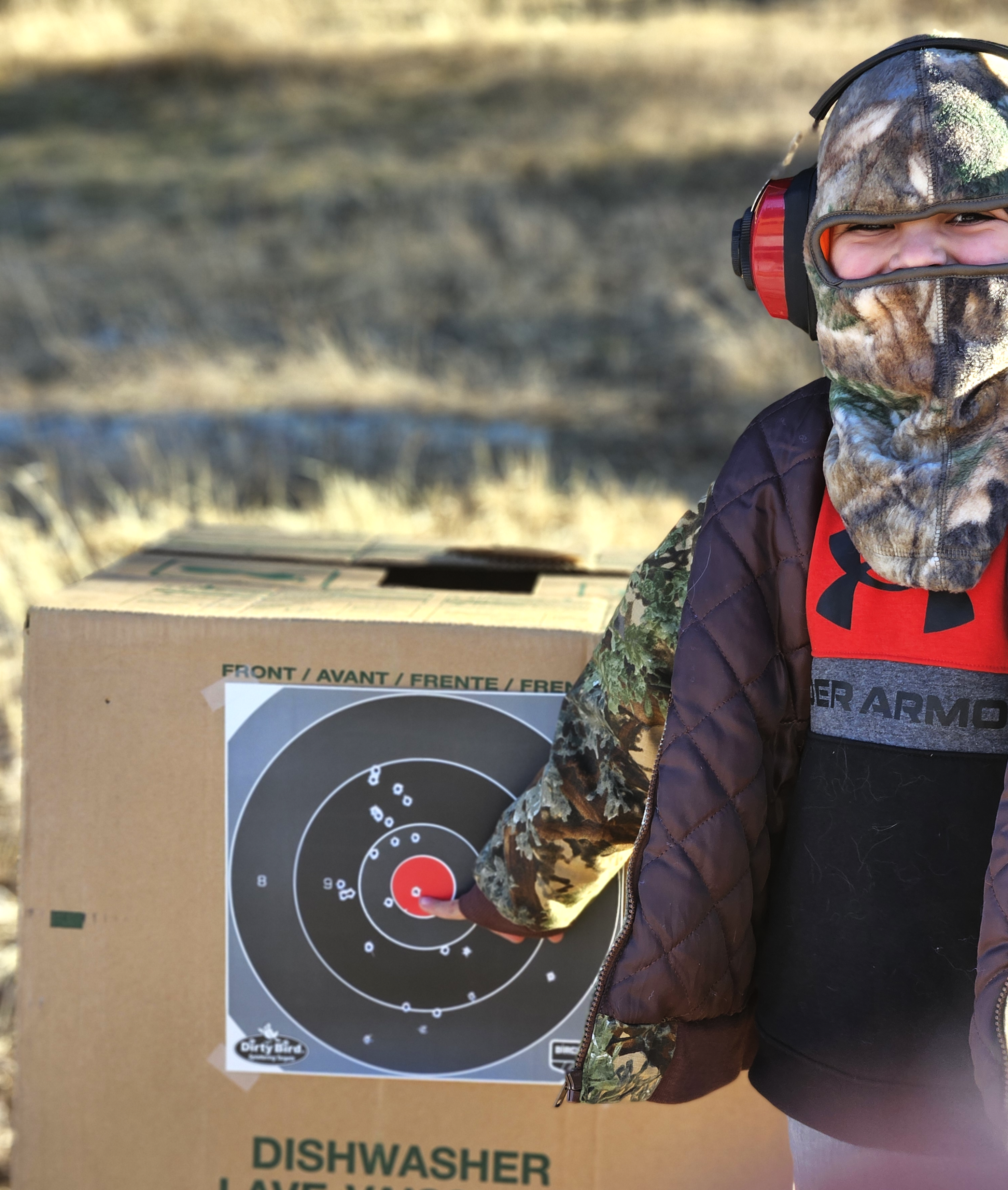 Child in camouflage jacket and face covering pointing at a target on a cardboard box outdoors during daytime.