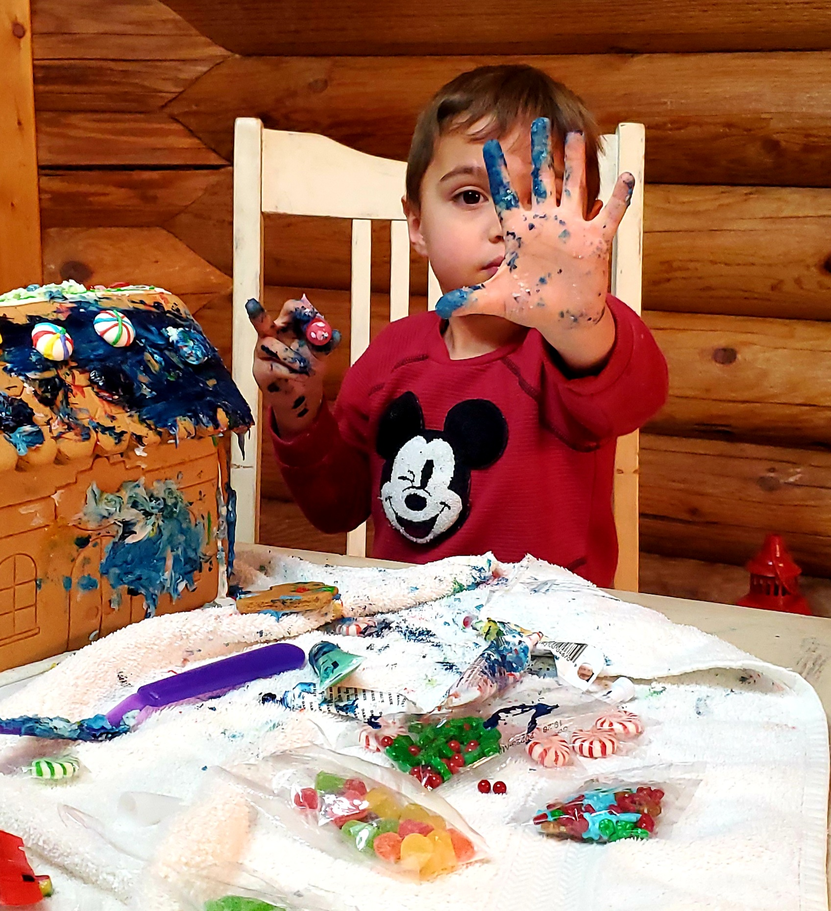 Young boy with painted hands and face, sitting at a table with candy and frosting, holding up his hand showing blue paint. Child wears red Mickey Mouse shirt, with cake and decoration supplies around.