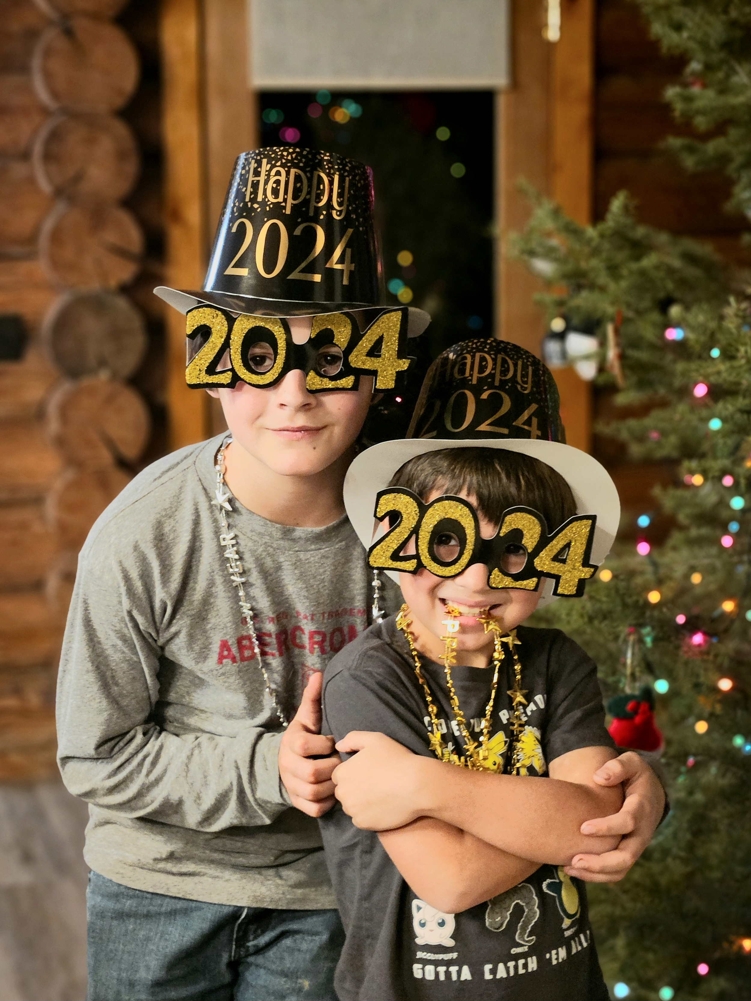 Two children celebrating New Year's Eve at 2024, wearing festive hats and glasses with '2024' and 'Happy 2024' written on them, standing in front of a Christmas tree with colorful lights, inside a wooden cabin.