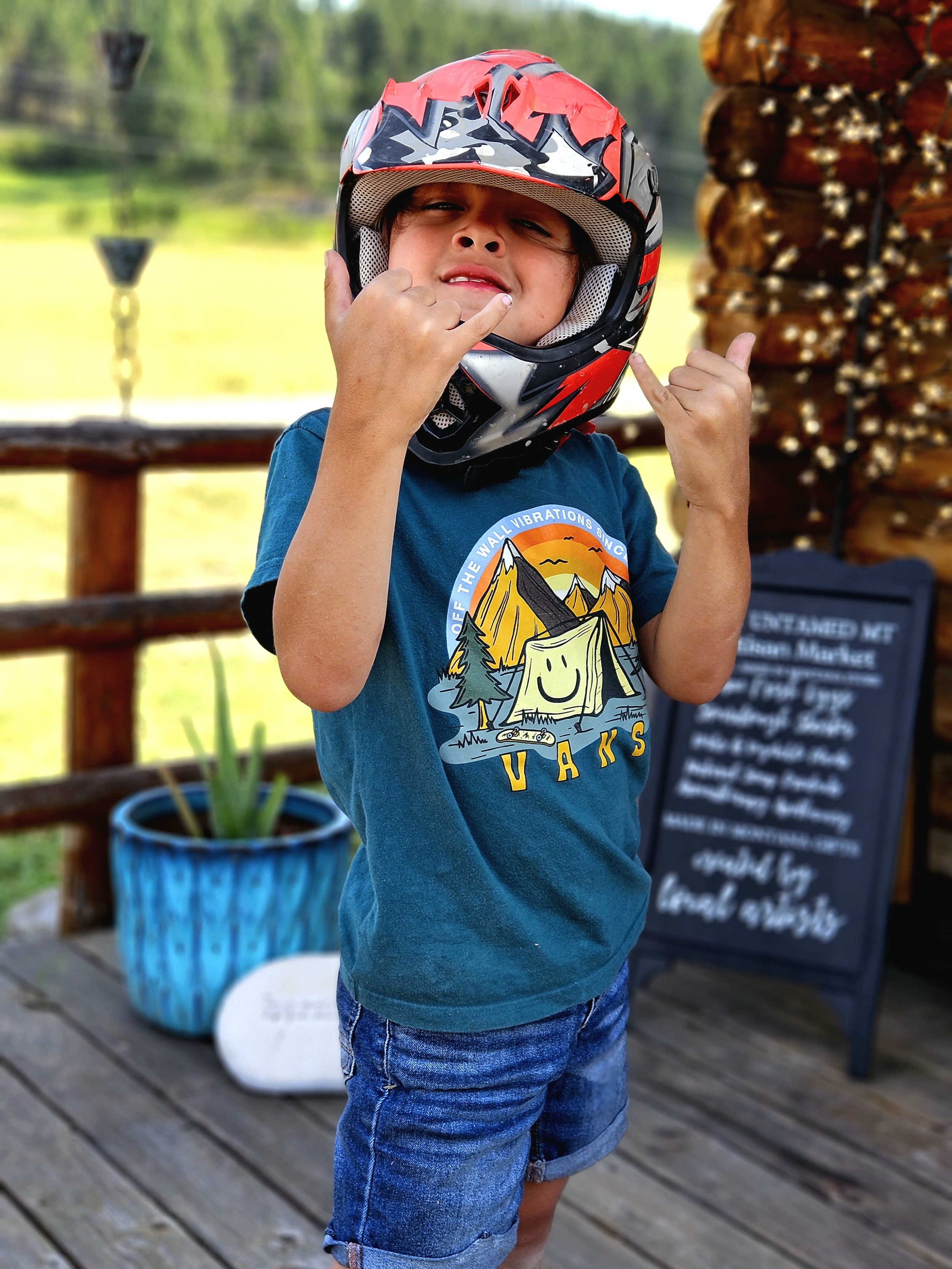 A young boy wearing a red and black motorcycle helmet making a shaka sign with both hands, standing on a wooden deck outdoors with a potted plant and a sign in the background.