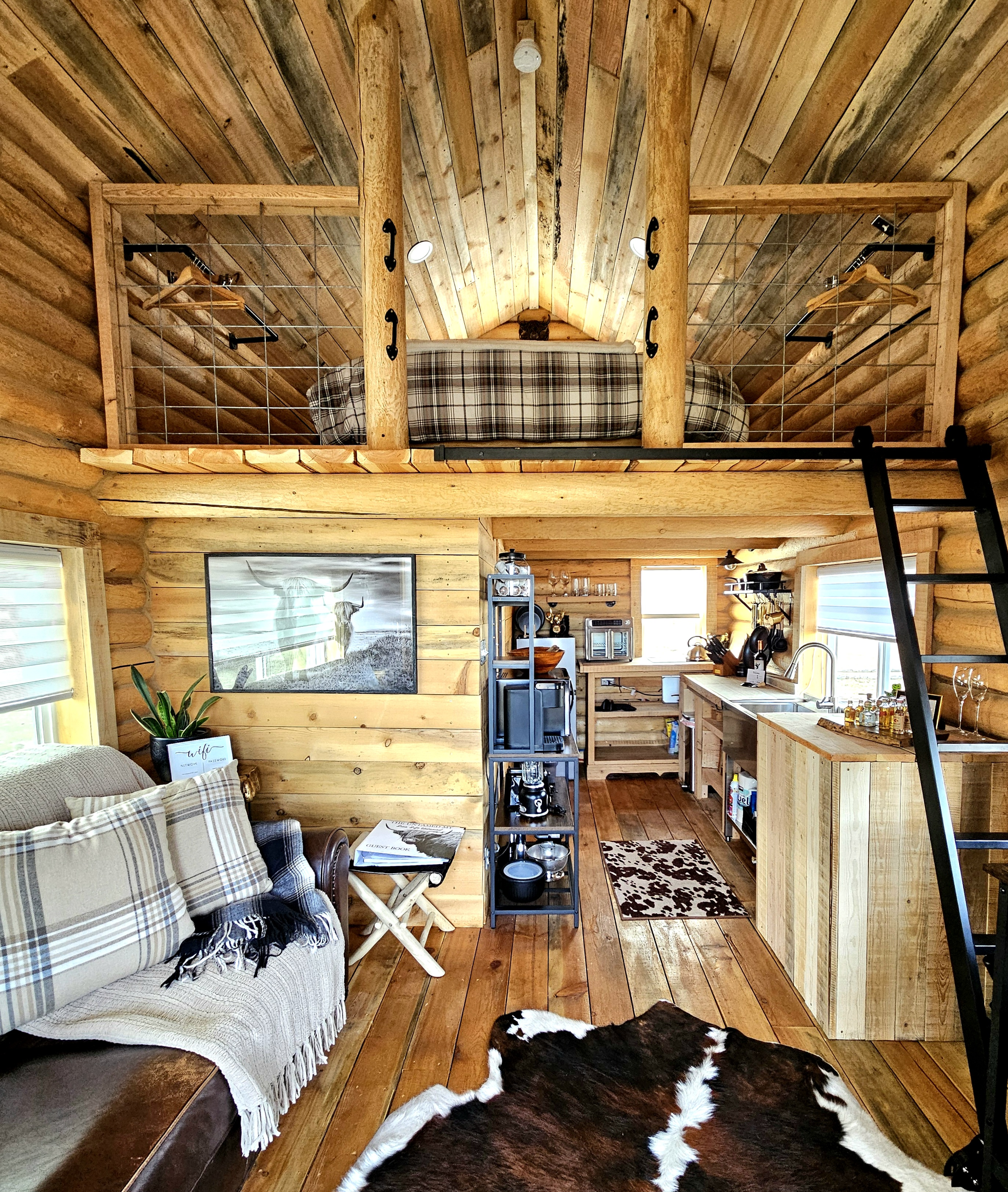 Interior view of a cozy log cabin with wood-paneled walls and ceiling, featuring a loft with a bed, a small living area with a couch and a cowhide rug, a compact kitchen, and a ladder leading to the loft.