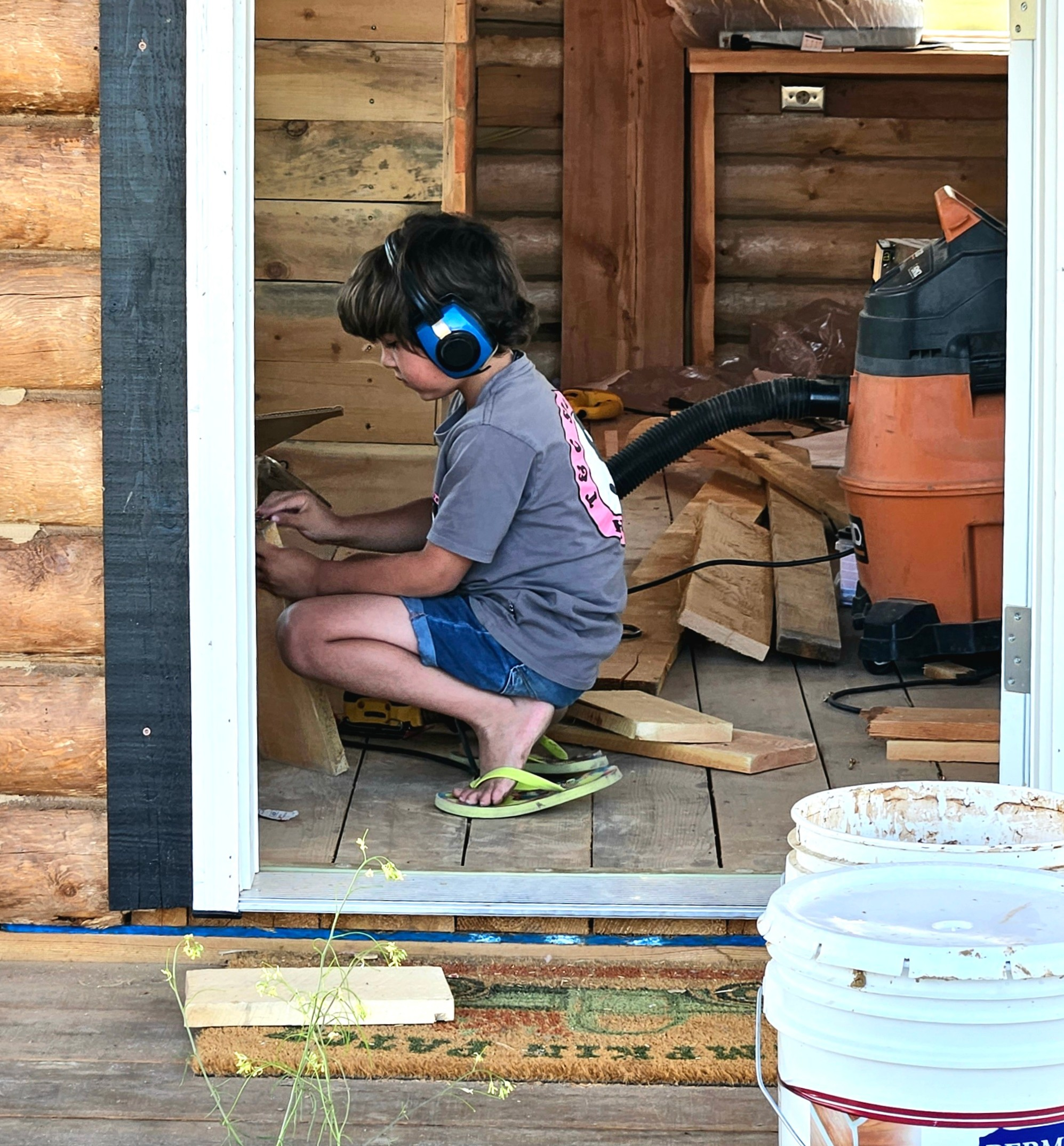 A boy kneeling inside a wooden shed, wearing headphones, working on a woodworking project with scattered pieces of wood, a shop vacuum, and buckets of supplies around him.