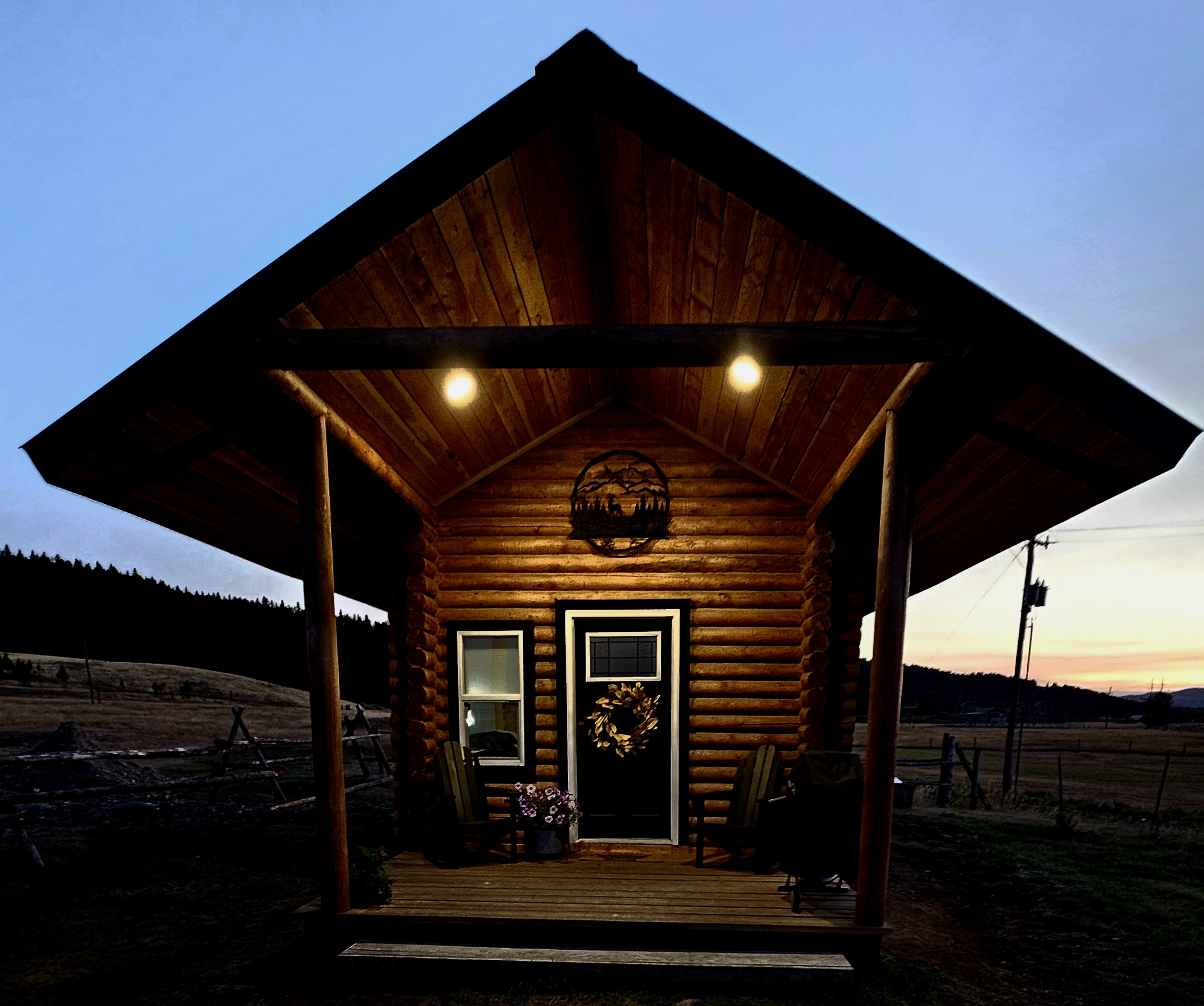 A small rustic log cabin with a front porch, two chairs, and a door decorated with a wreath, illuminated by two porch lights at dusk, with open fields and a sunset in the background.