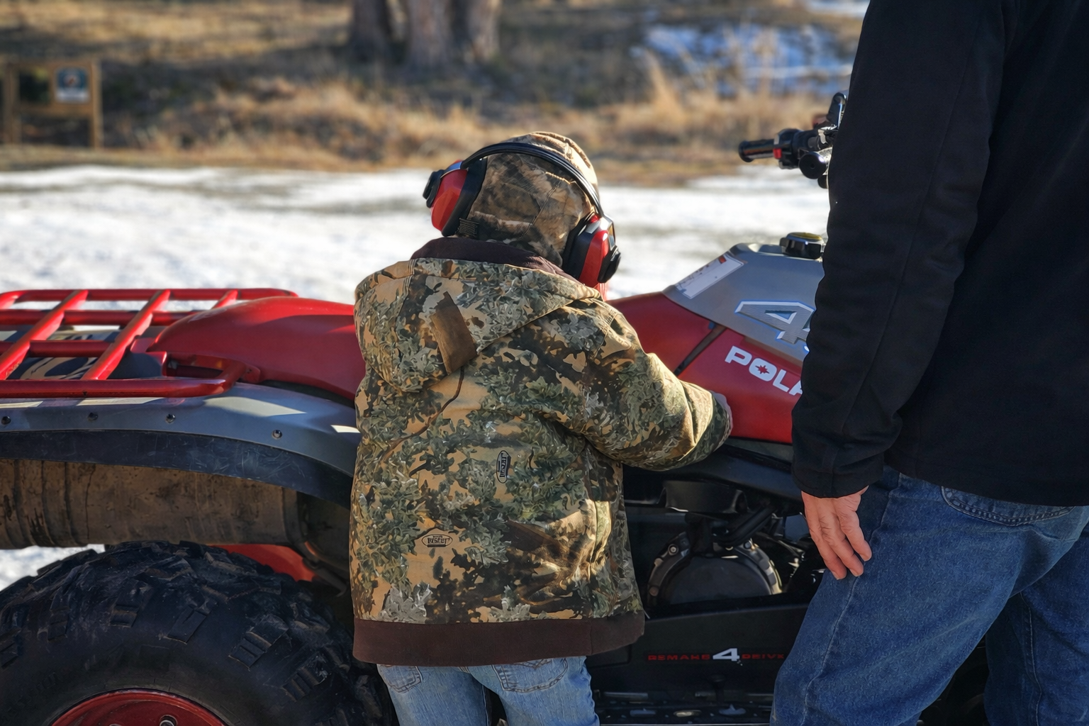 Child wearing camouflage jacket and earmuffs inspecting a red and gray ATV, with an adult standing nearby.