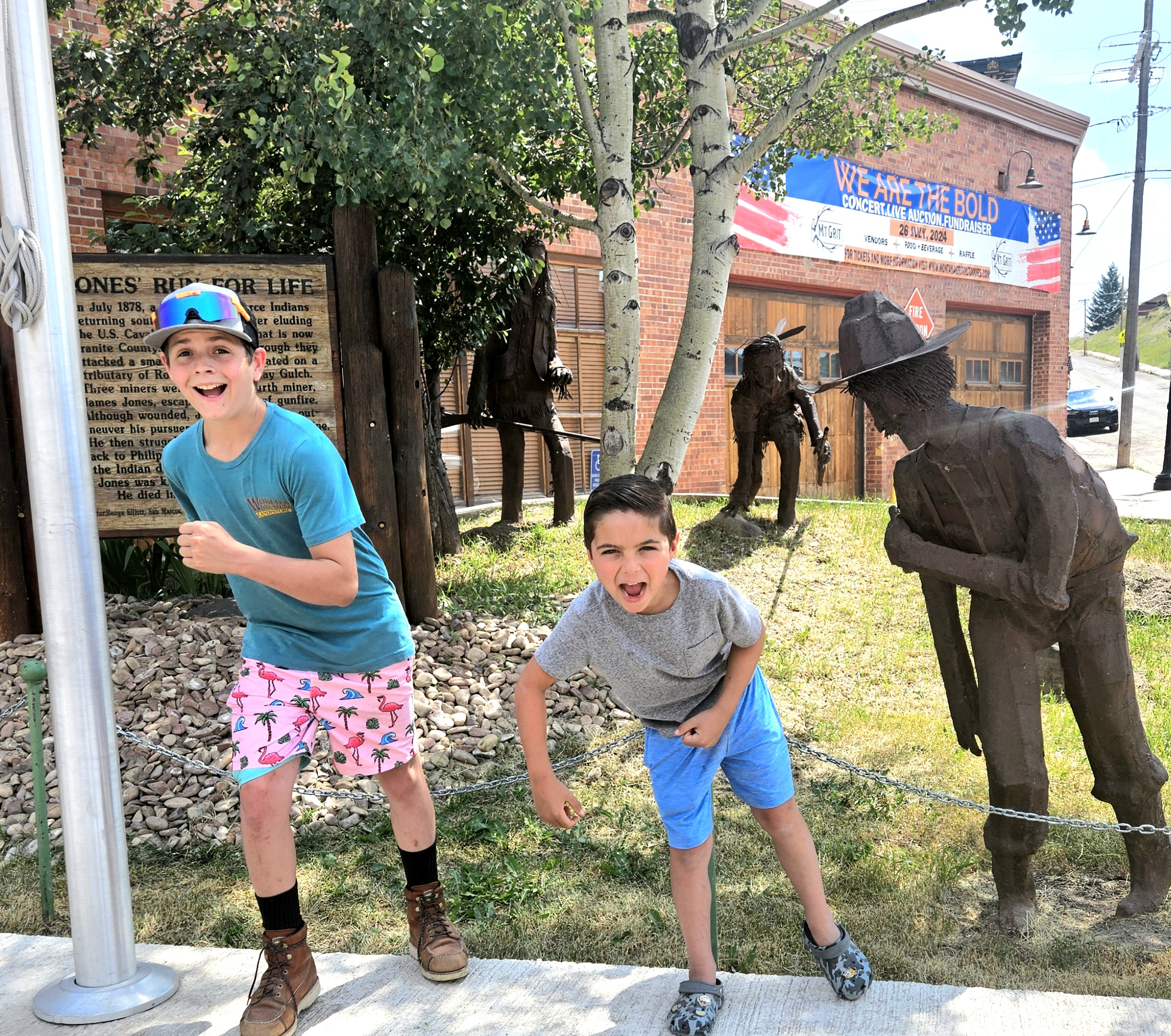 Two boys with playful expressions posing in front of bronze statues of historical figures on a small patch of grass lined with a chain, with a brick building and a blue banner in the background.