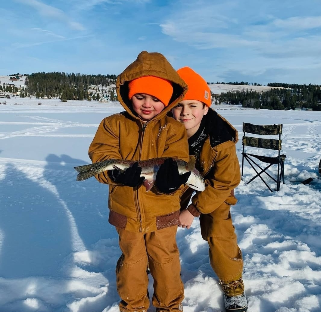 Two boys in winter clothing holding a large fish on a snowy lake with a forested shoreline in the background.