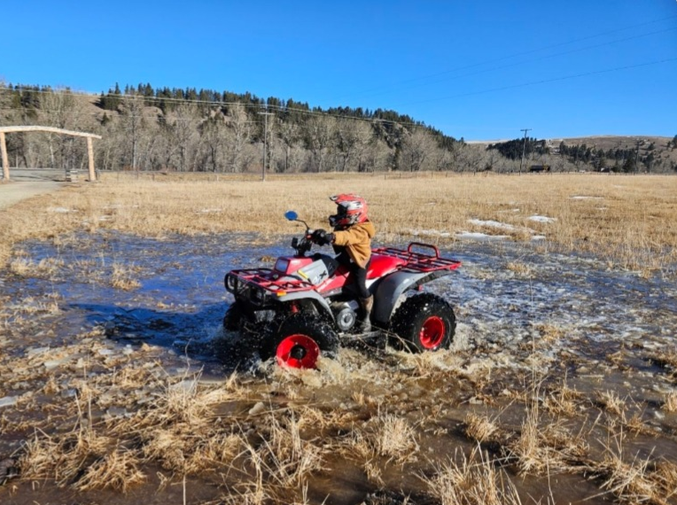 Child riding a red and black all-terrain vehicle through a flooded field on a sunny day.