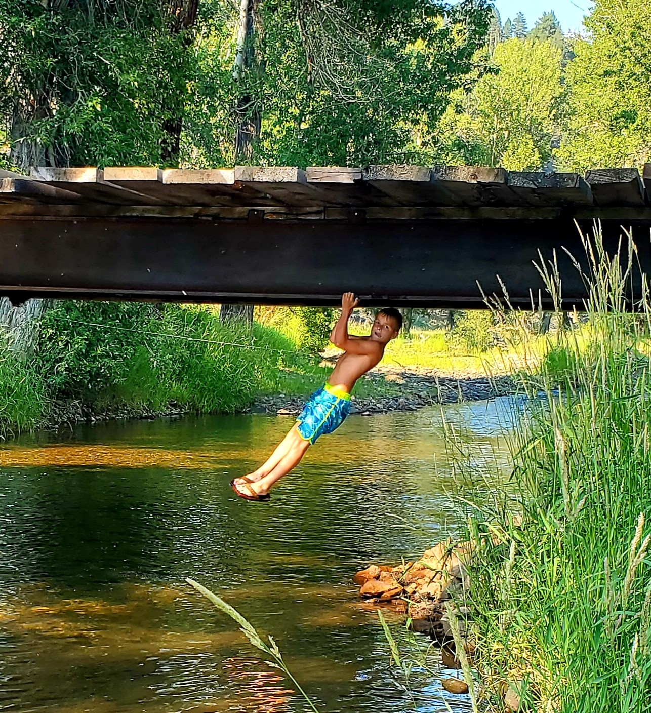 A young boy in blue swim trunks hanging from a bridge over a creek, with his body partially under the bridge and the rest of his body hanging hanging in the water.