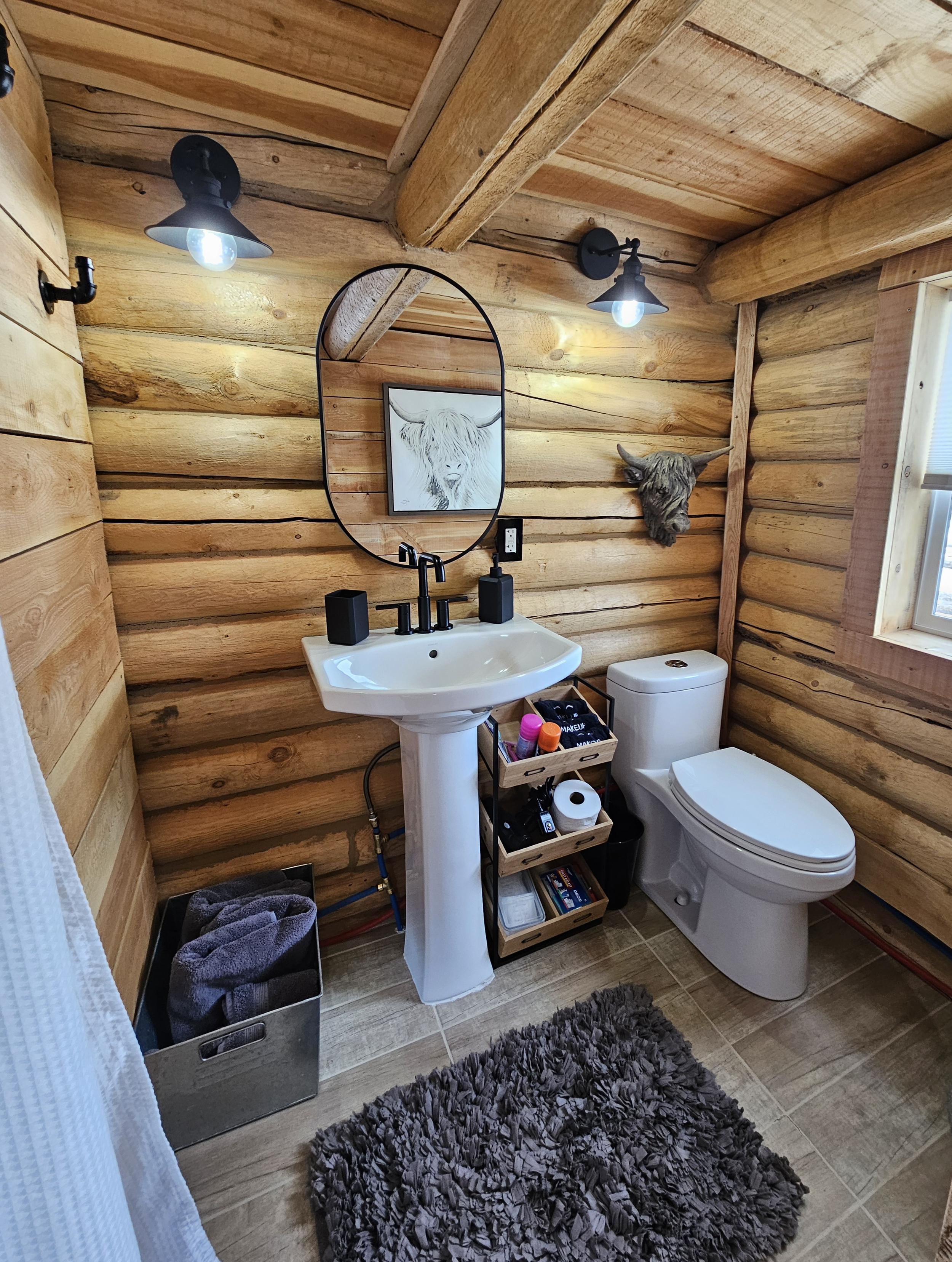 Bathroom with wooden log walls and ceiling, a white pedestal sink with black fixtures, a round mirror, a toilet, a black metal storage cart with toiletries, a gray shaggy rug, and decorative animal skull and drawing on the wall.