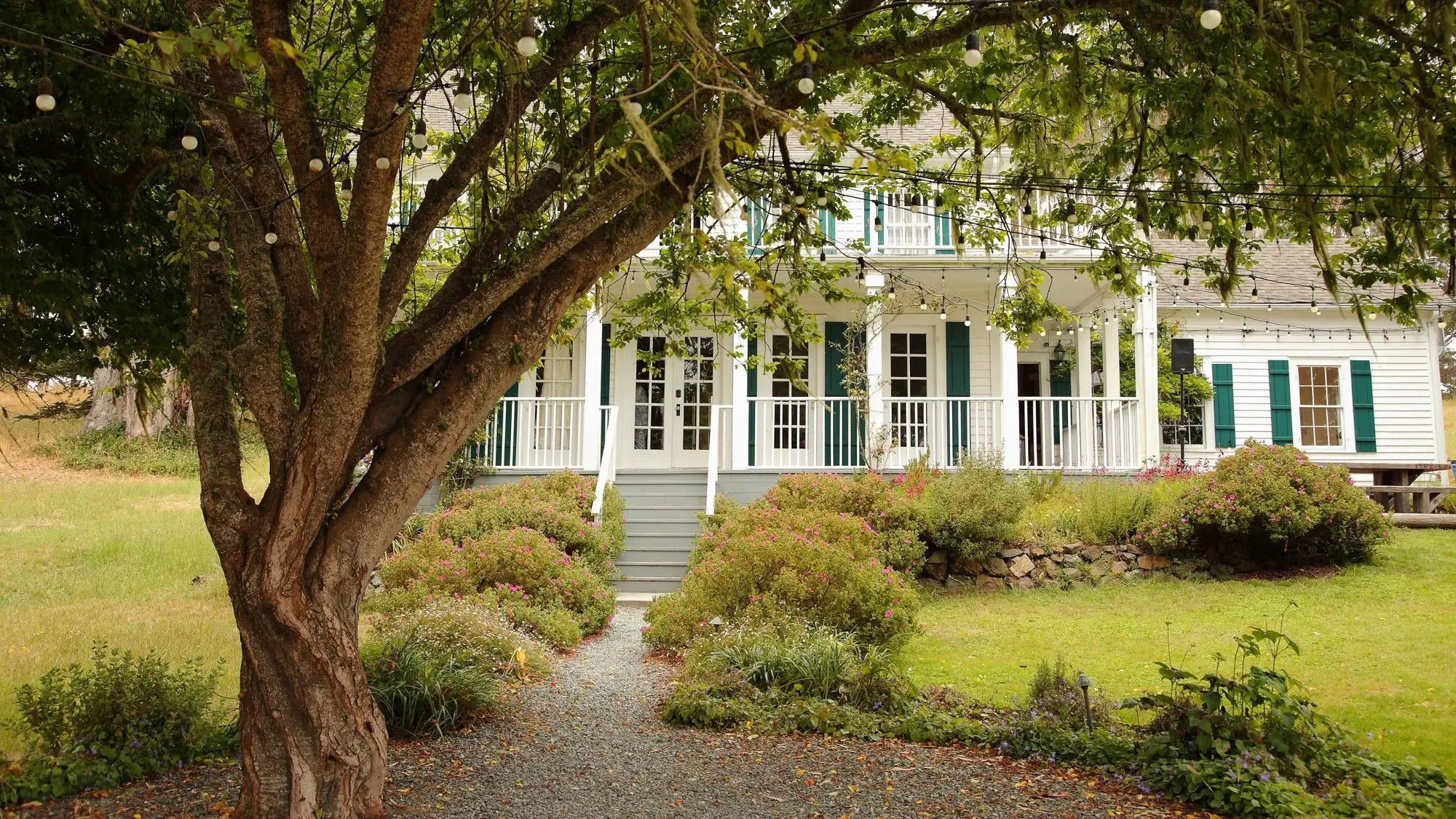 Charming white two-story house with green window shutters, front porch, and surrounding garden; large tree and string lights in foreground.