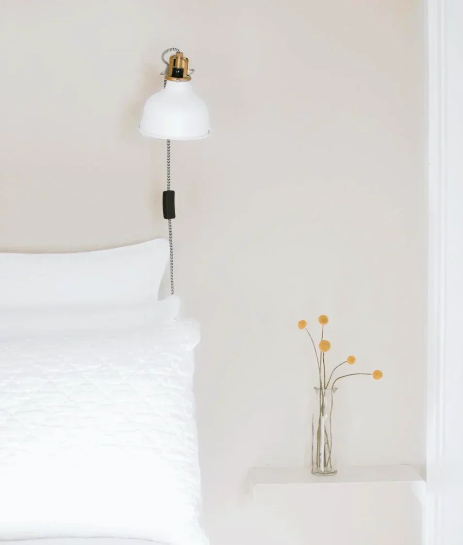 White bed under a wall lamp with a small vase of yellow flowers
