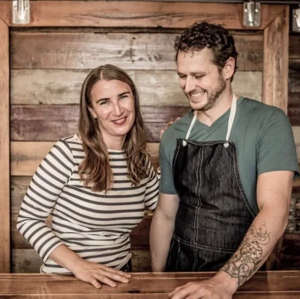 A woman in a striped shirt and a man in an apron smiling at a wooden counter