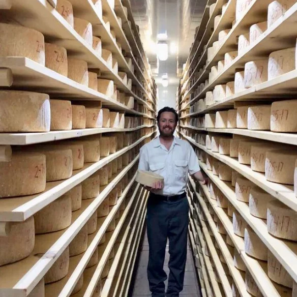 A cheesemaker standing in an aisle between tall shelves of aging cheese wheels