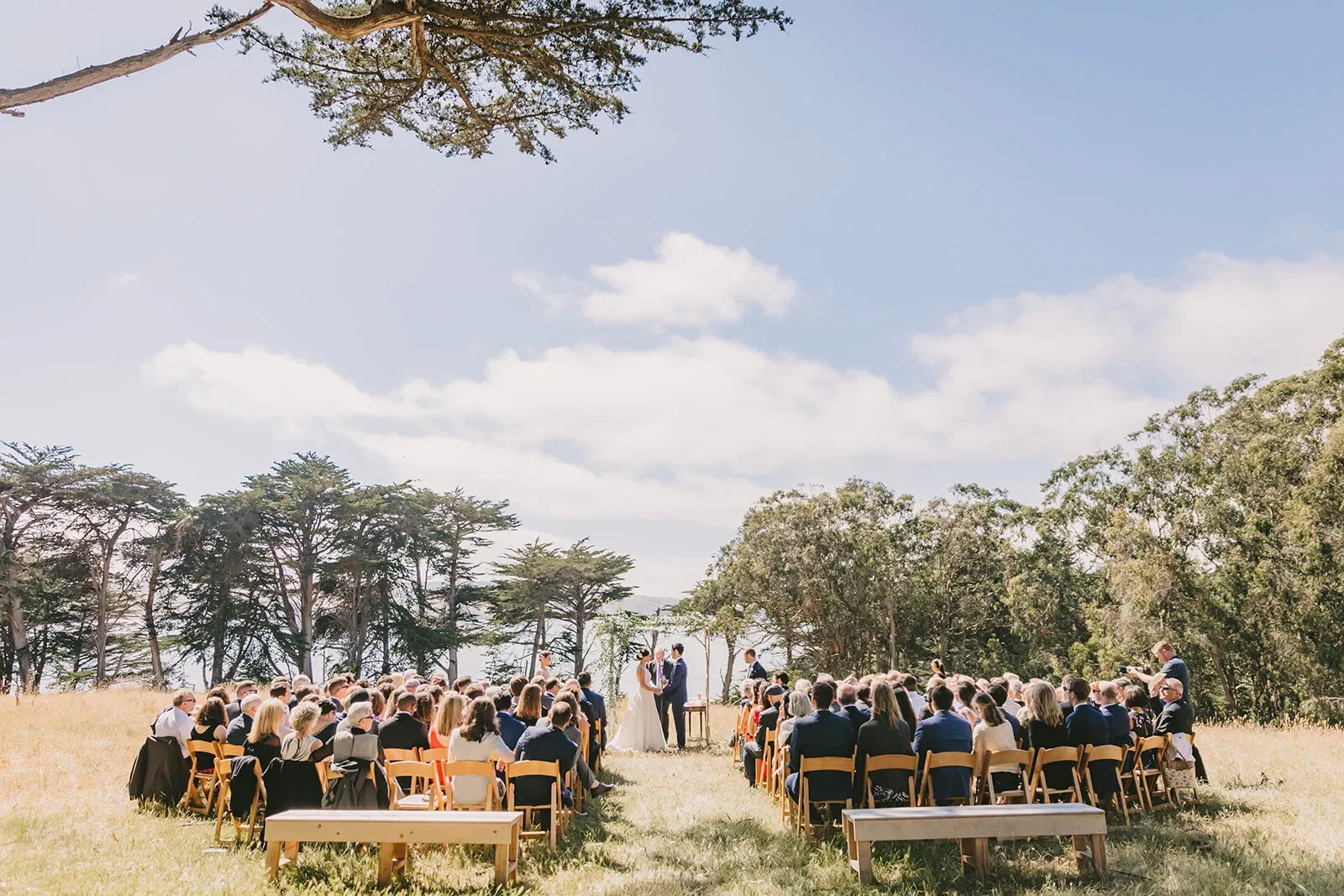 Outdoor wedding ceremony in a dry grass meadow, guests on wooden chairs facing the couple, cypress trees behind with a blue sky and white puffy clouds.