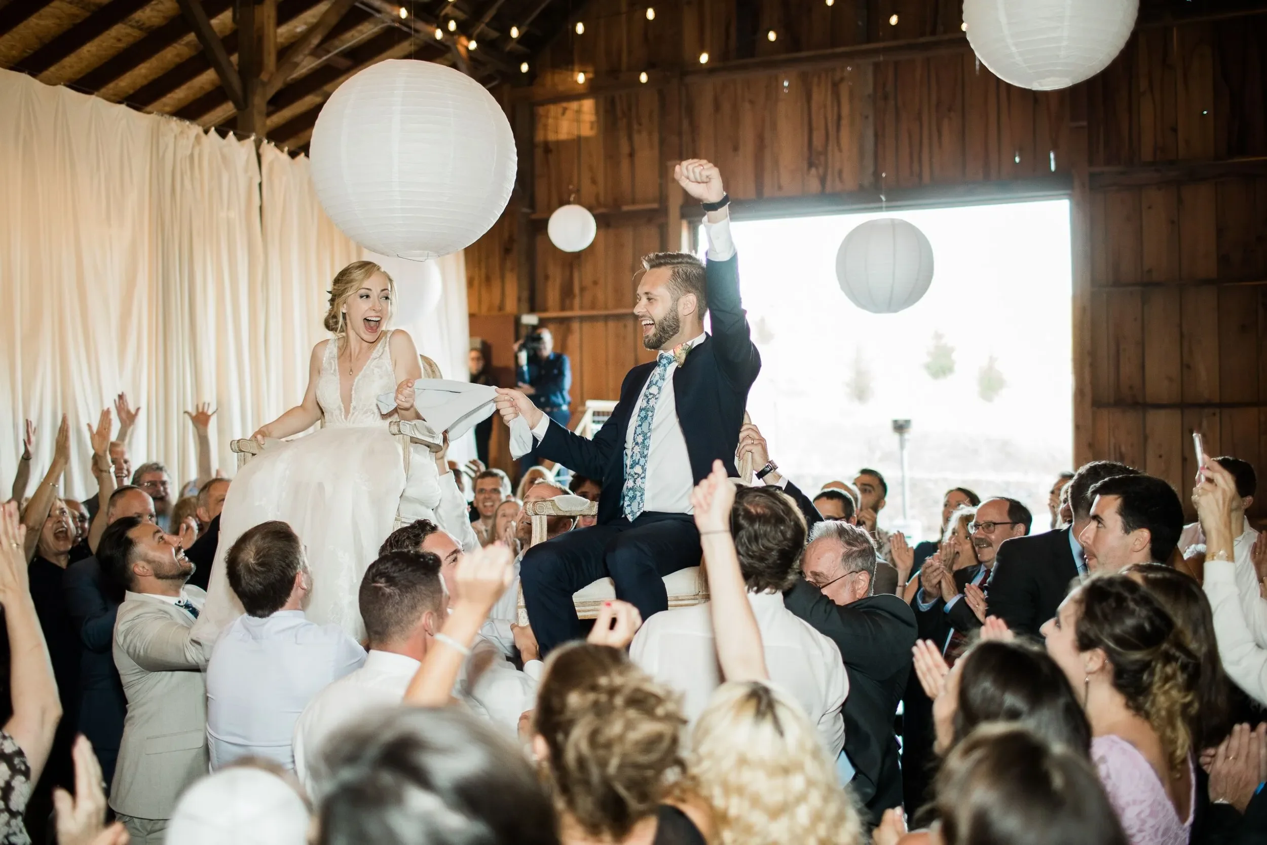 Bride and groom being lifted on chairs in a lively wedding celebration indoors with guests clapping and cheering, decorated with paper lanterns.