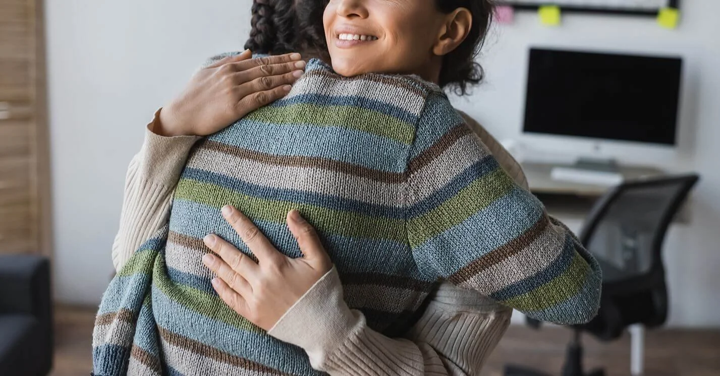 Two people hugging in an office, one smiling with curly hair, wearing a beige sweater, the other with braided hair, wearing a colorful striped sweater.