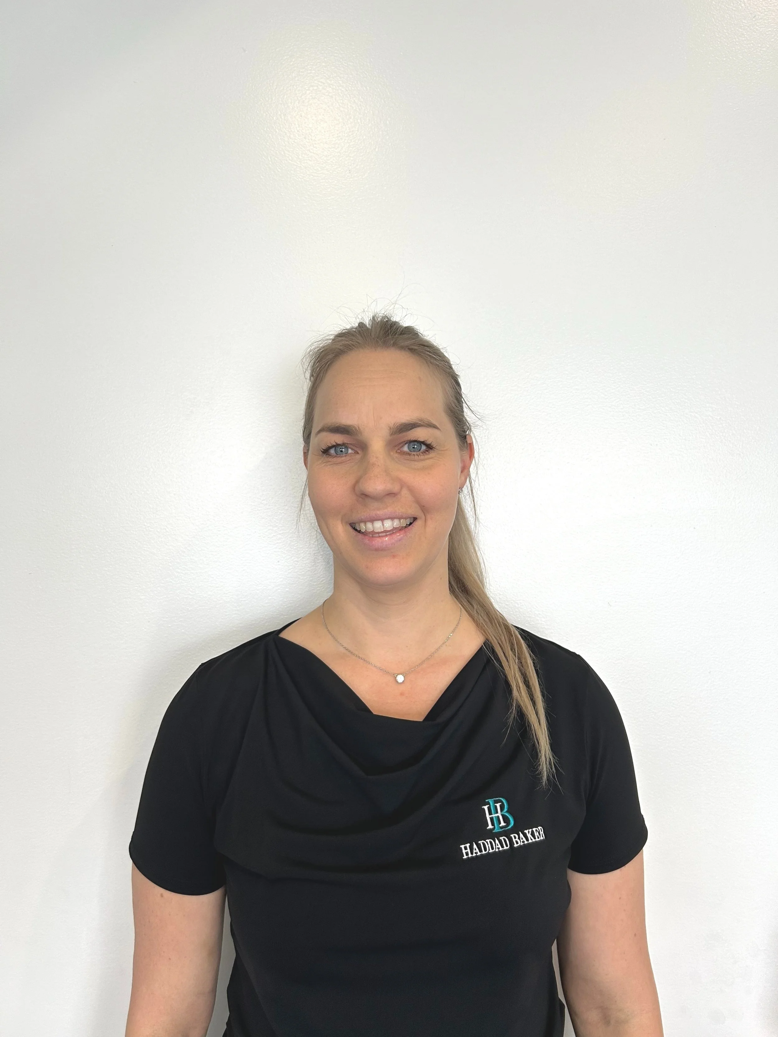 A smiling woman with blonde hair pulled back, wearing a black shirt with 'Haddad Baker' logo, standing against a plain white wall.