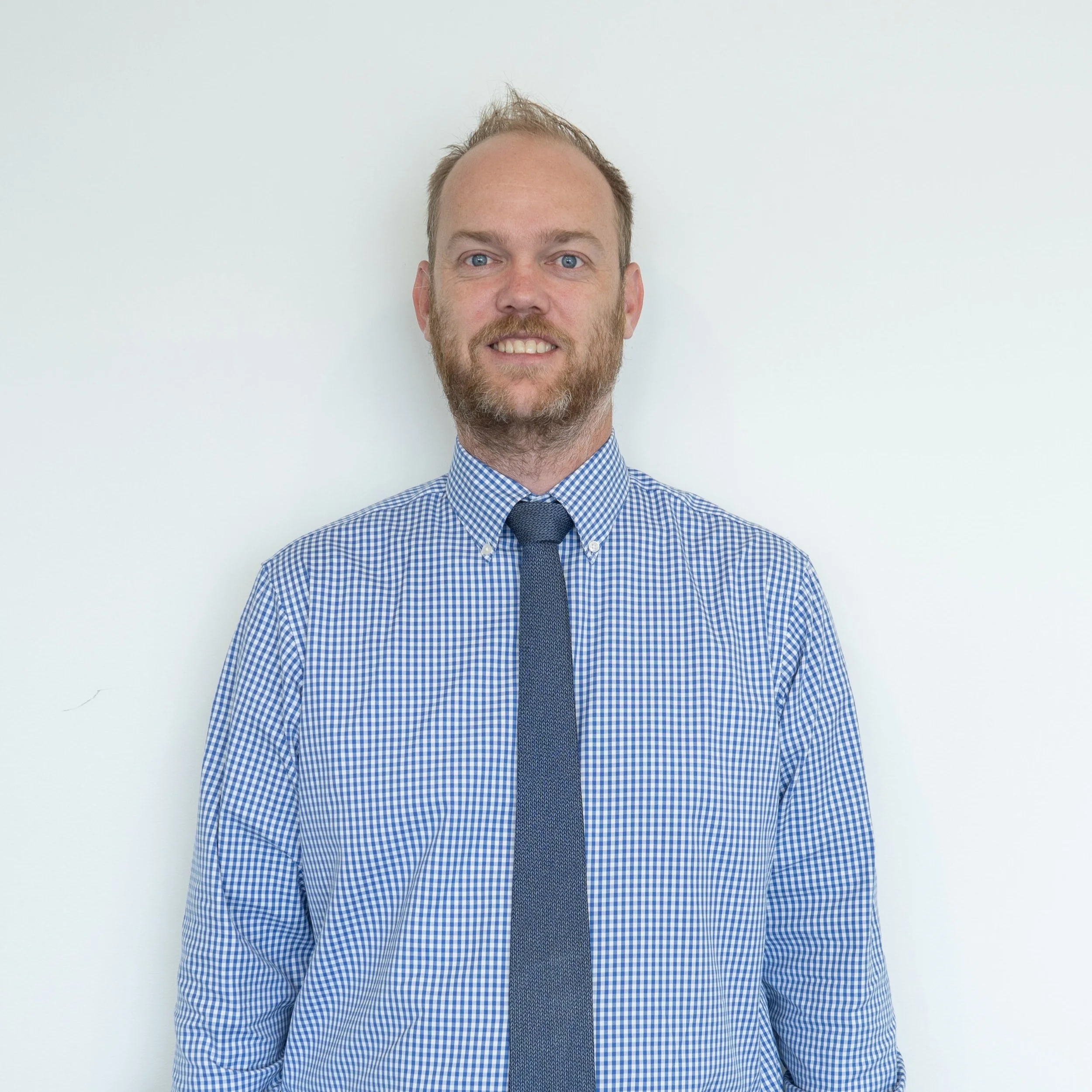 A man with a beard and light brown hair wearing a blue checkered dress shirt and a navy blue tie, standing against a plain white wall, smiling at the camera.