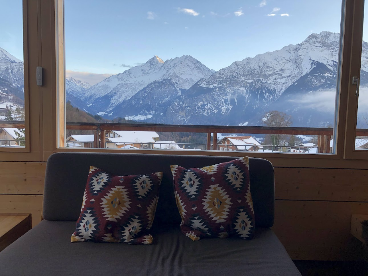 Sofa with patterned cushions placed in front of a large window framing a wide view of snow-covered mountains and alpine village rooftops.
