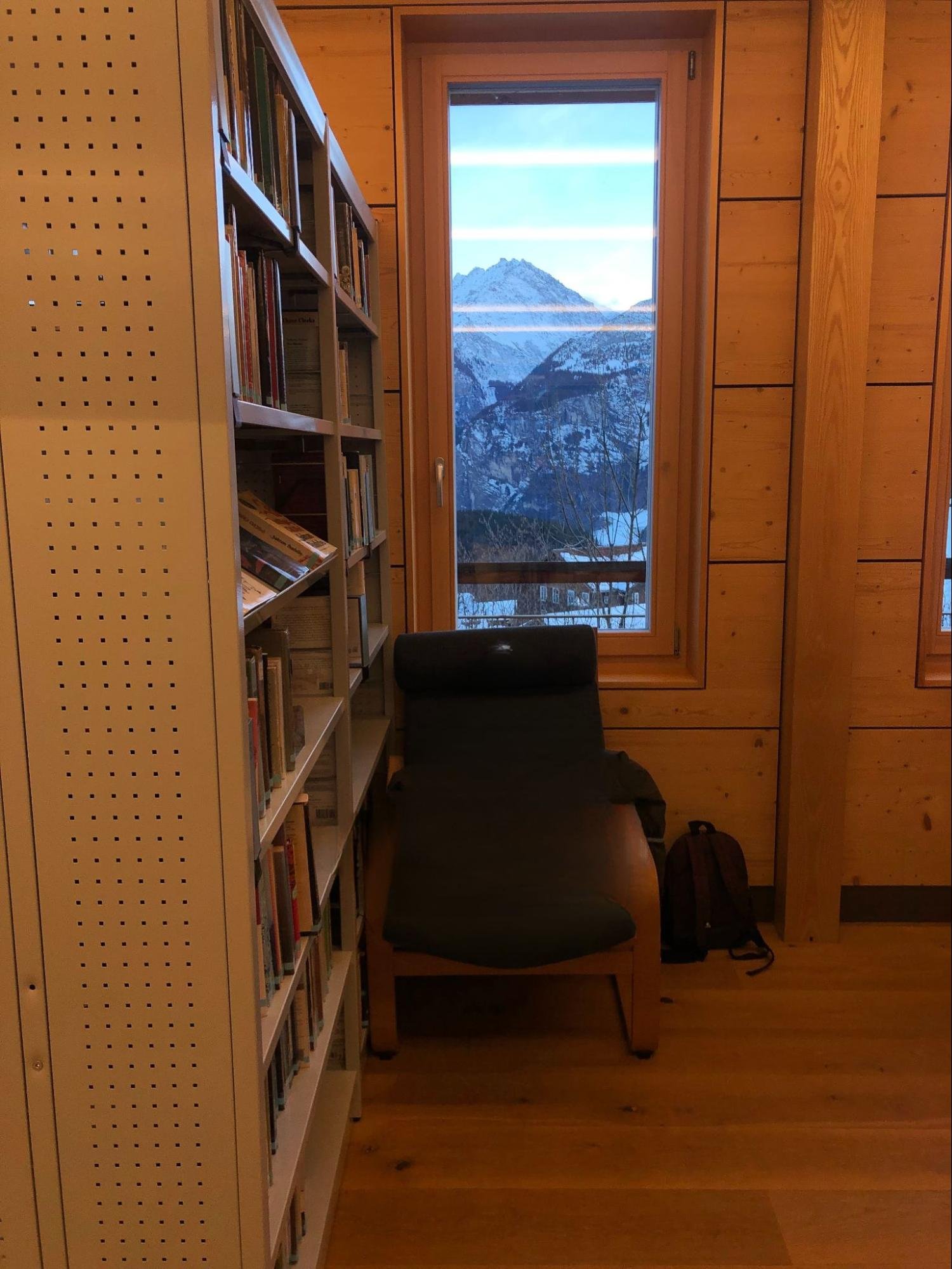 Cozy wooden library corner with bookshelves and a lounge chair facing a tall window overlooking snow-covered alpine peaks.