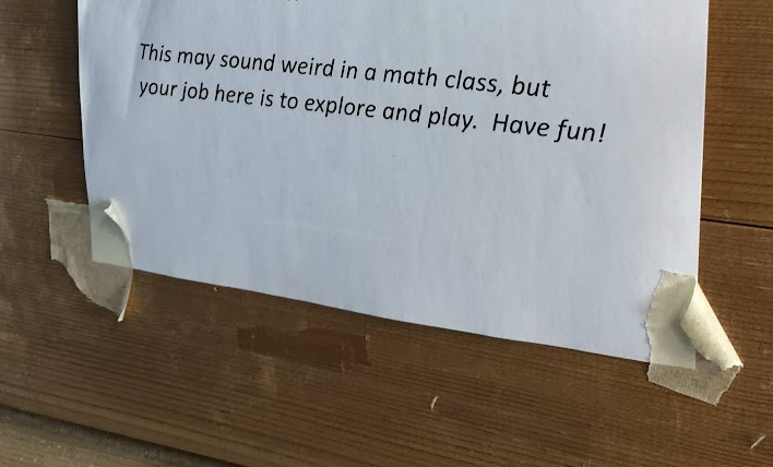 Printed classroom sign taped to a wooden wall reading: “This may sound weird in a math class, but your job here is to explore and play. Have fun!”