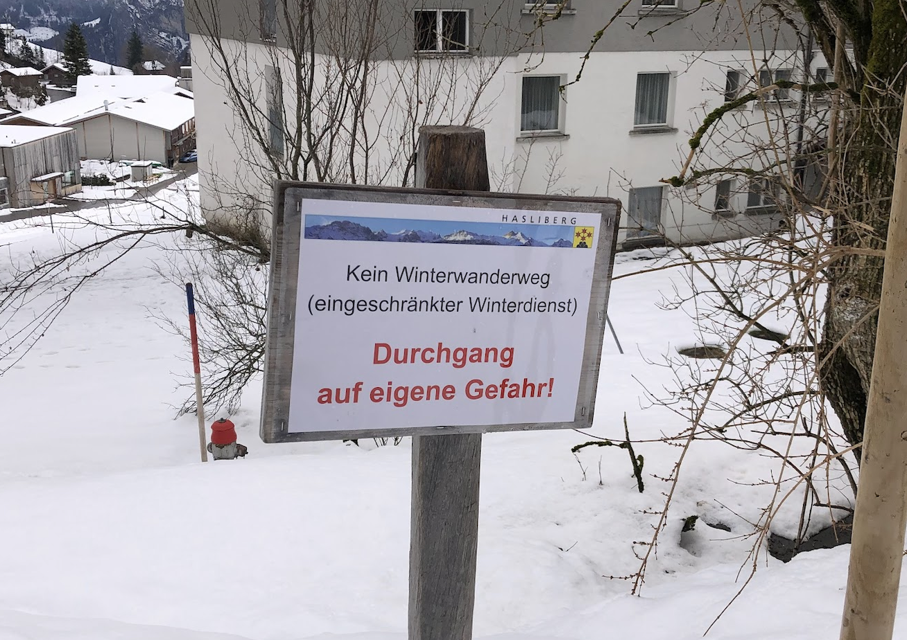 Sign in the snow in Hasliberg reading “Kein Winterwanderweg (eingeschränkter Winterdienst) – Durchgang auf eigene Gefahr!” in front of residential buildings.