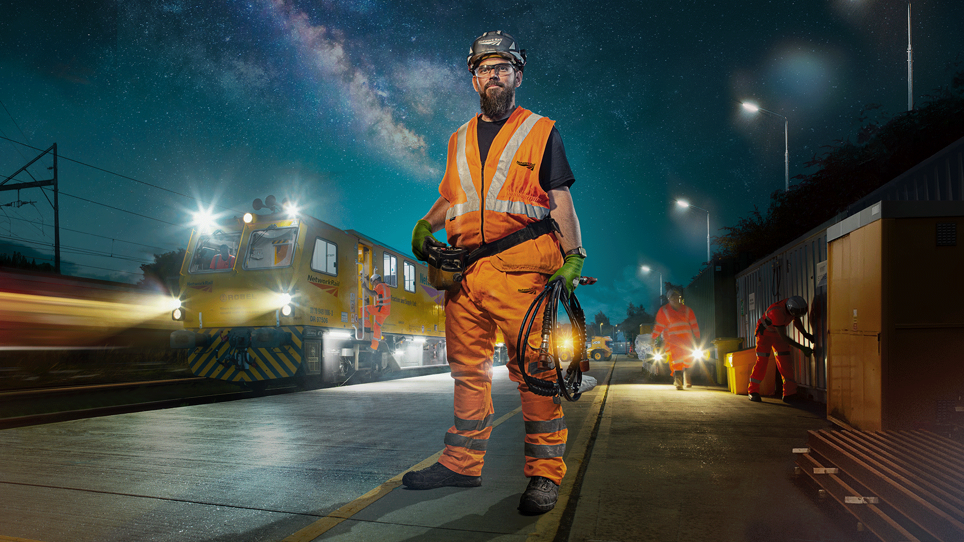 Jamie, one of Network Rail's frontline workers, stands proudly and heroically next to a bright yellow maintenance train in a Network Rail depot