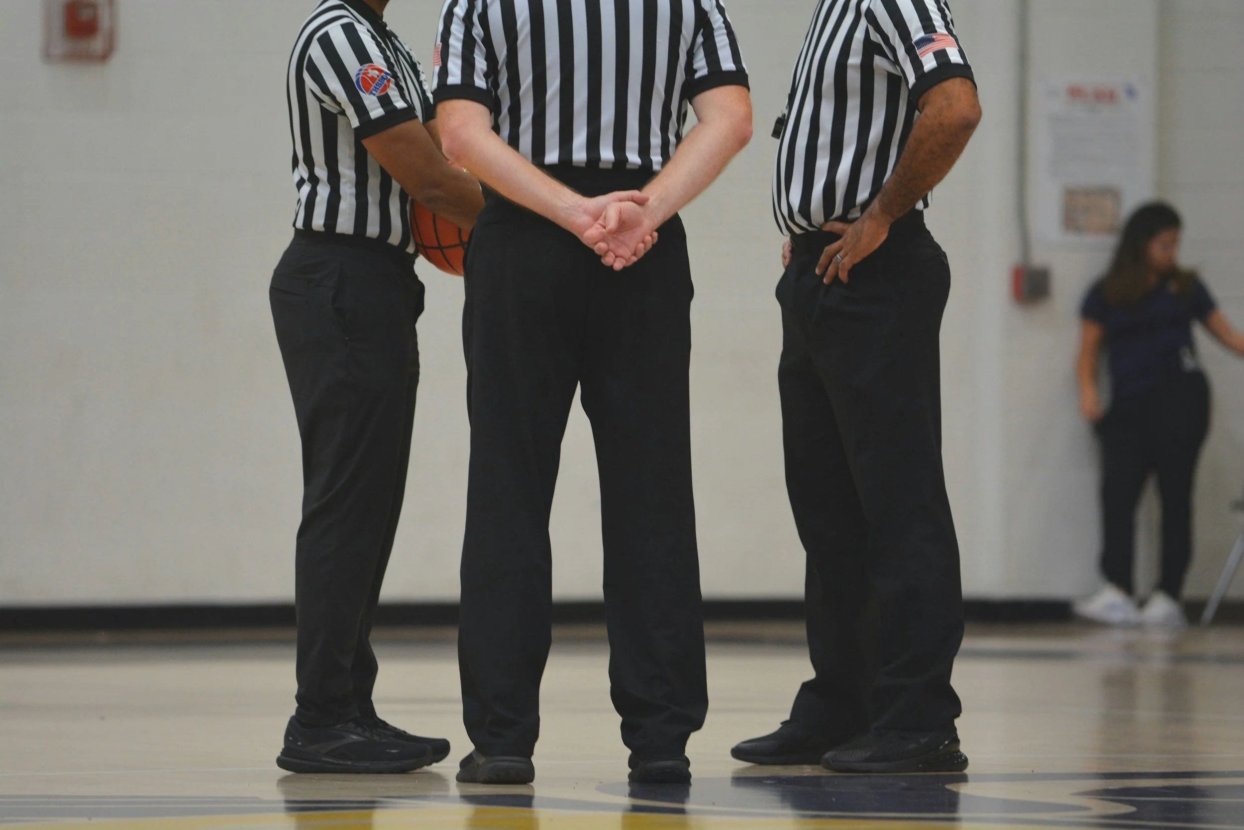 Three referees in striped shirts and black pants standing on a gymnasium floor, engaged in a discussion, with a person in the background.