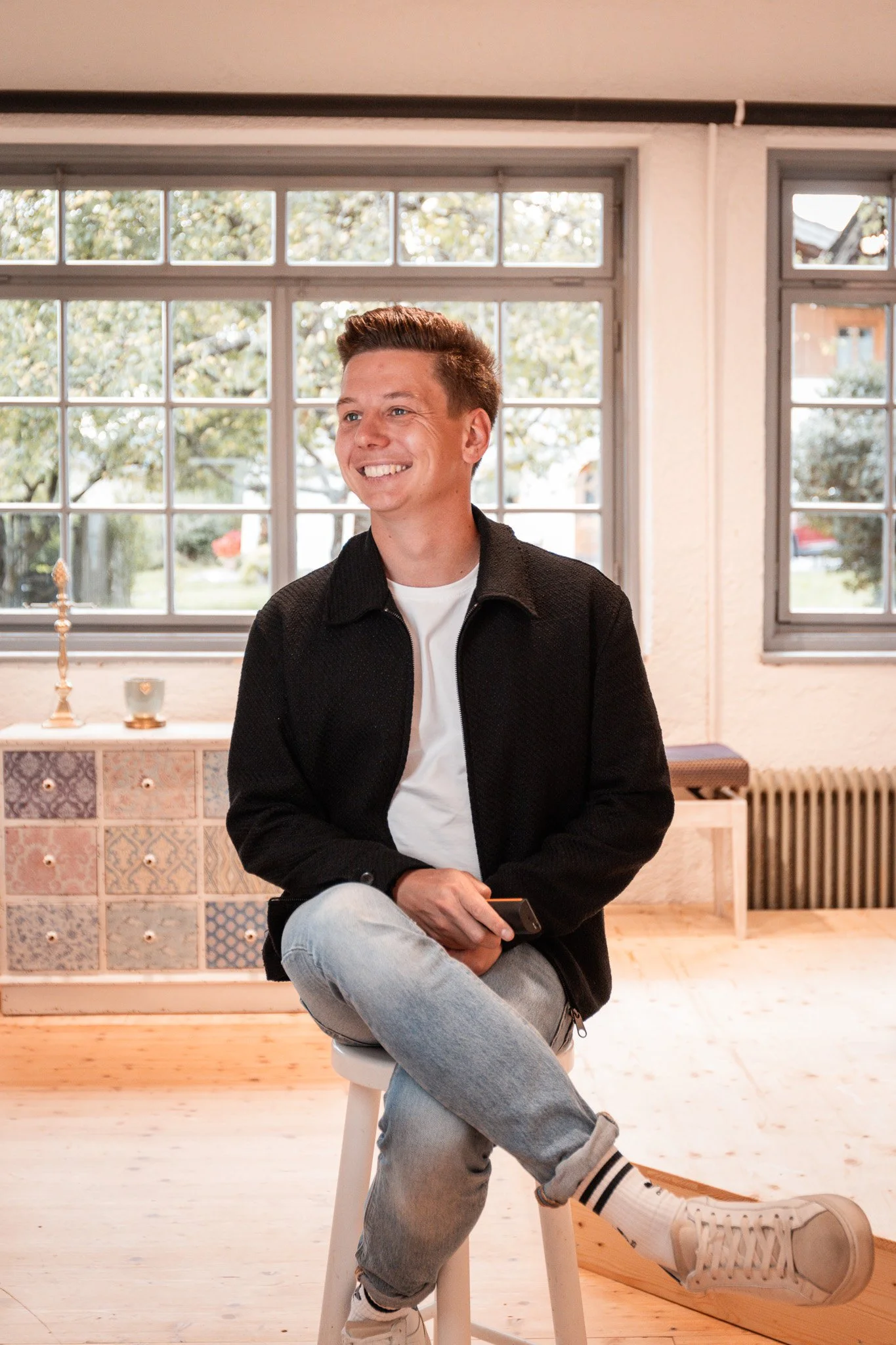 Sebastian Schmidt sitzt auf einem Hocker, lächelt und hält ein Smartphone in der Hand, im Hintergrund große Fenster mit Blick nach draußen.
