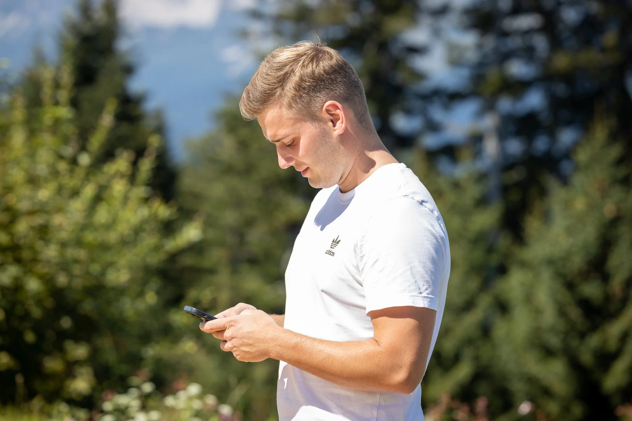 Junger Mann liest auf einem Smartphone im Freien, umgeben von Bäumen und Natur, bei sonnigem Wetter.