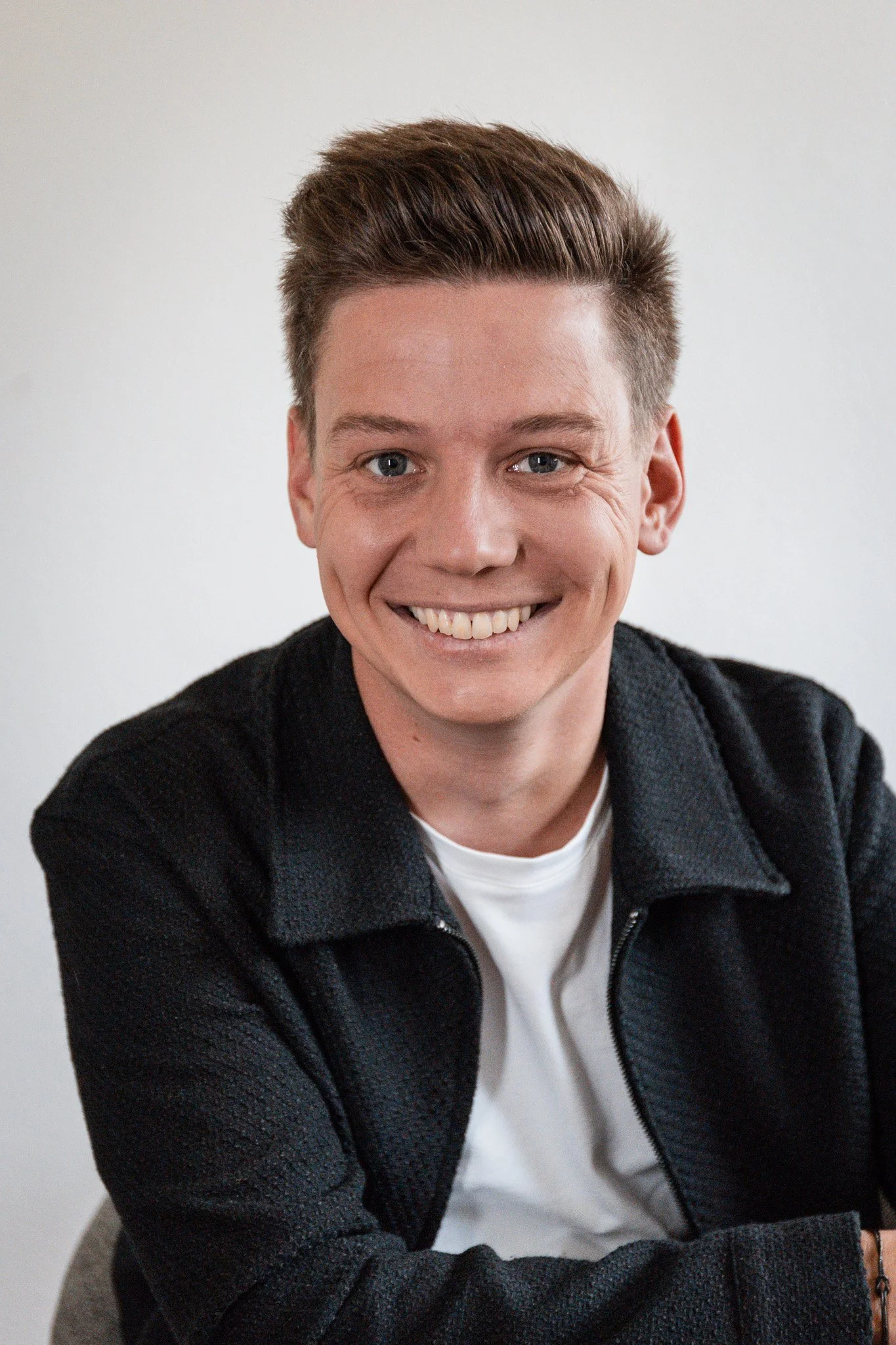 Sebastian Schmidt mit braunen Haaren, trägt schwarzen Pullover und weißes T-Shirt, lächelnd.