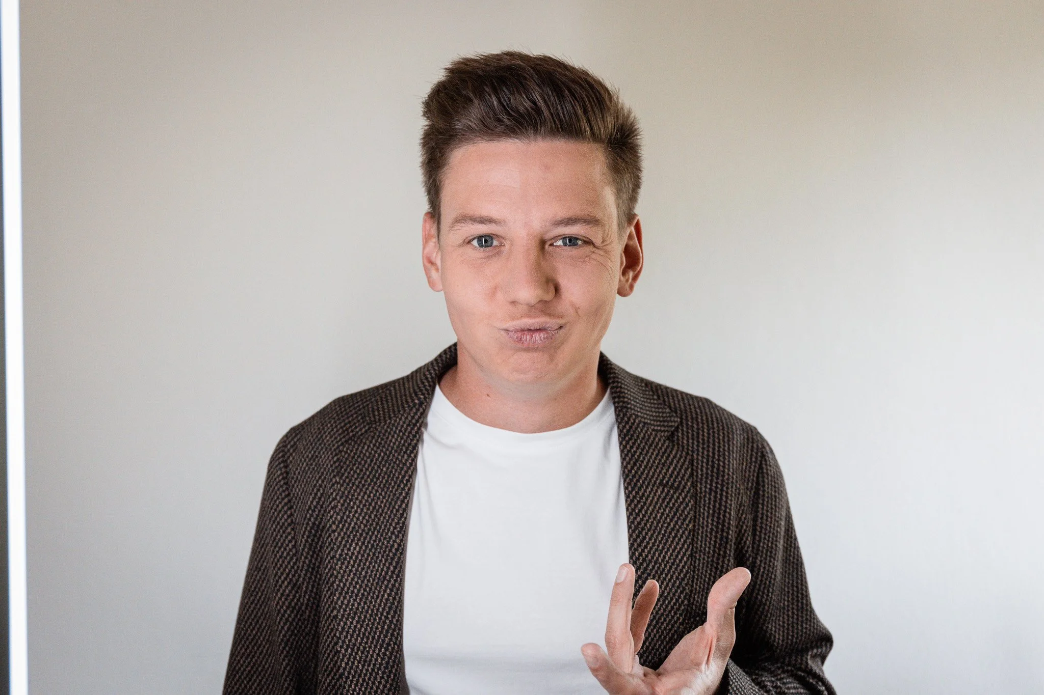 Sebastian Schmidt macht eine lustige Grimasse, trägt einen weißen T-Shirt und ein kariertes Jackett, vor hellem Hintergrund.
